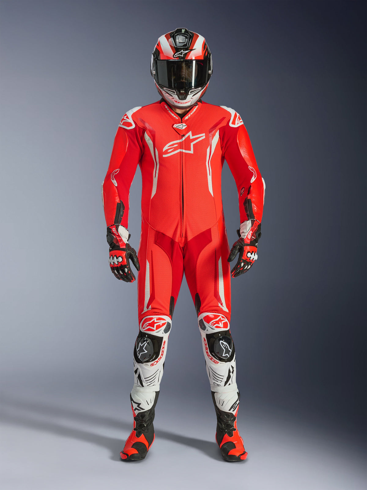 Pilote portant la combinaison en cuir une pièce Alpinestars GP Tech V5 en Rouge Fluo, Rouge Vif et Blanc, avec casque de course, gants et bottes assortis. Combinaison de piste professionnelle avec silhouette aérodynamique, logos Alpinestars blancs et sliders de protection intégrés.