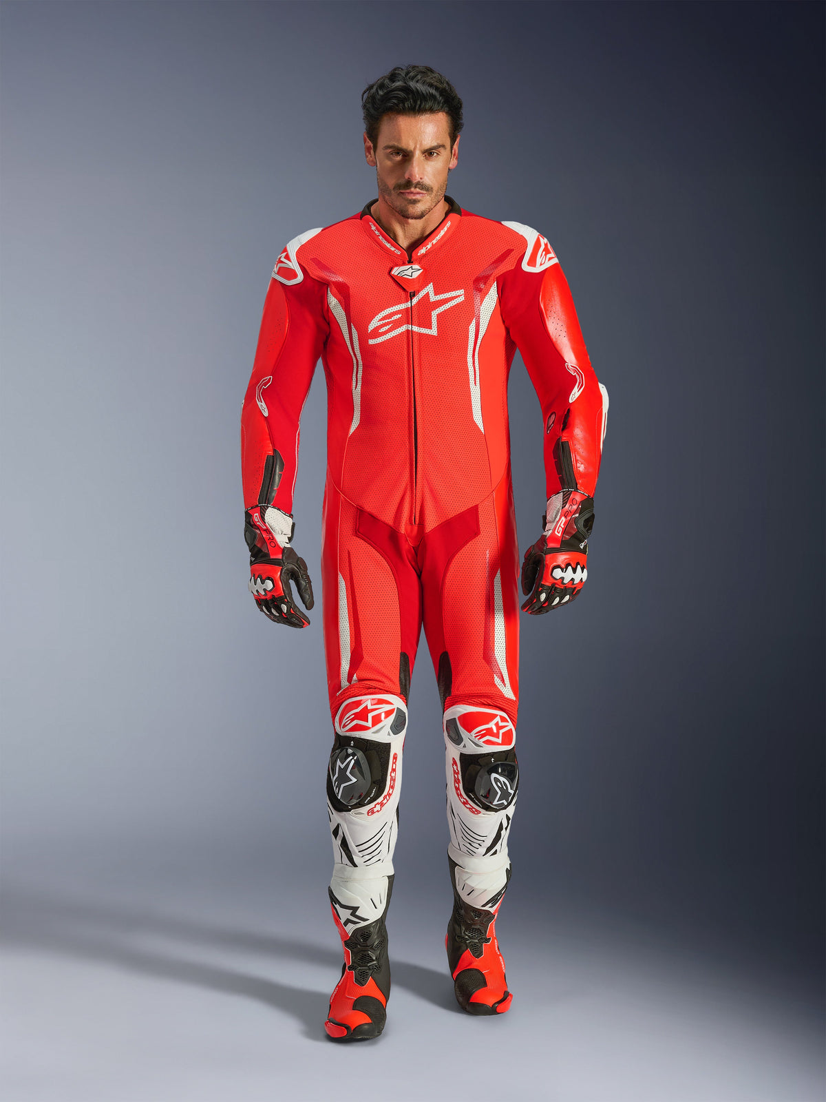 Alpinestars GP Tech V5, combinaison de course une pièce en cuir, combinaison professionnelle rouge fluo et rouge vif avec des accents blancs, portée par un pilote avec des bottes et des gants assortis, dotée d'une bosse aérodynamique, d'une construction en cuir de première qualité et de protections avancées pour la performance sur circuit