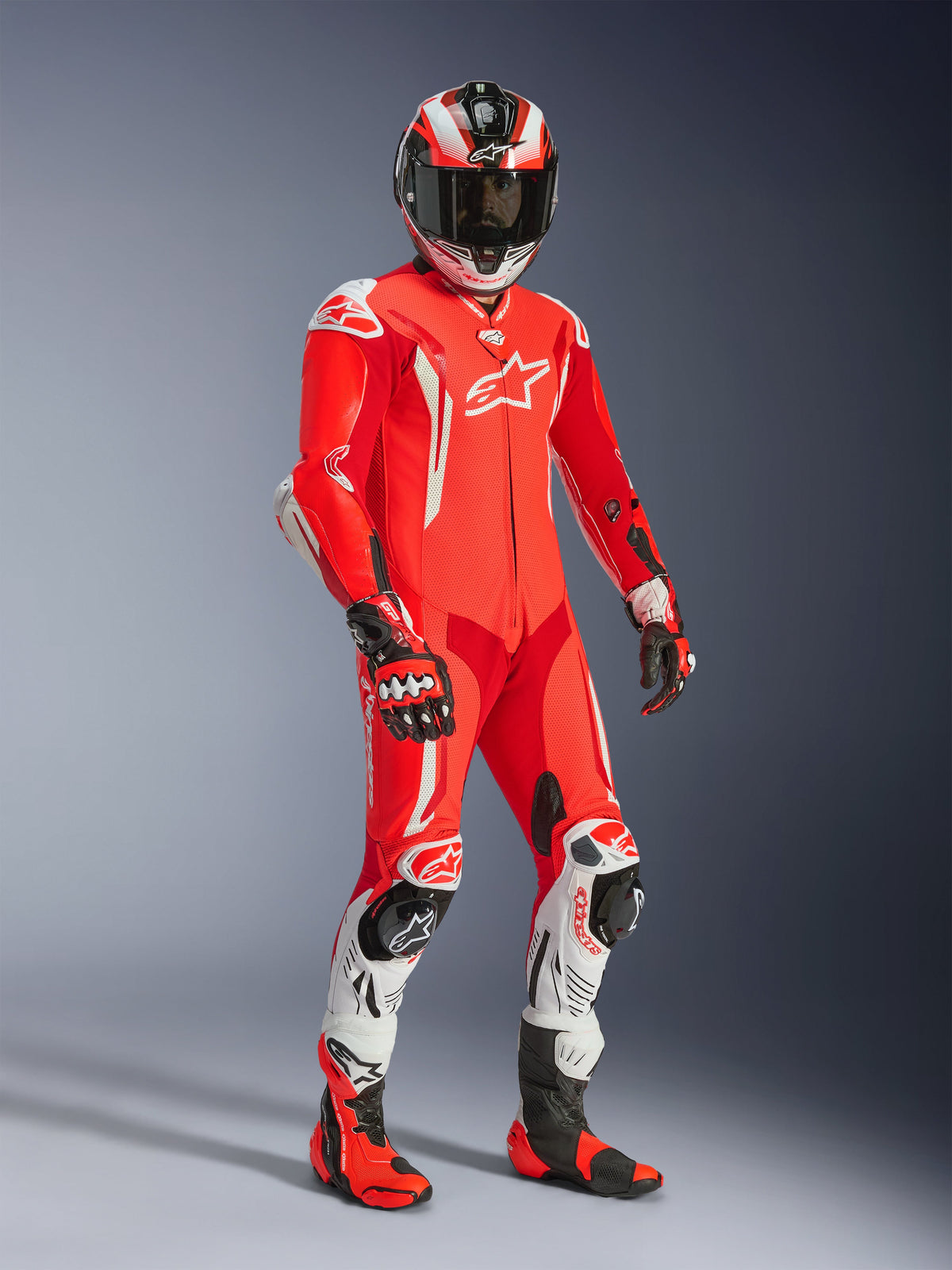 Pilote de moto portant la combinaison en cuir Alpinestars GP Tech V5 une pièce, rouge fluo et rouge vif avec des accents blancs, comprenant un casque, des gants et des bottes de course assortis, équipement de piste professionnel avec cuir perforé et sliders de protection