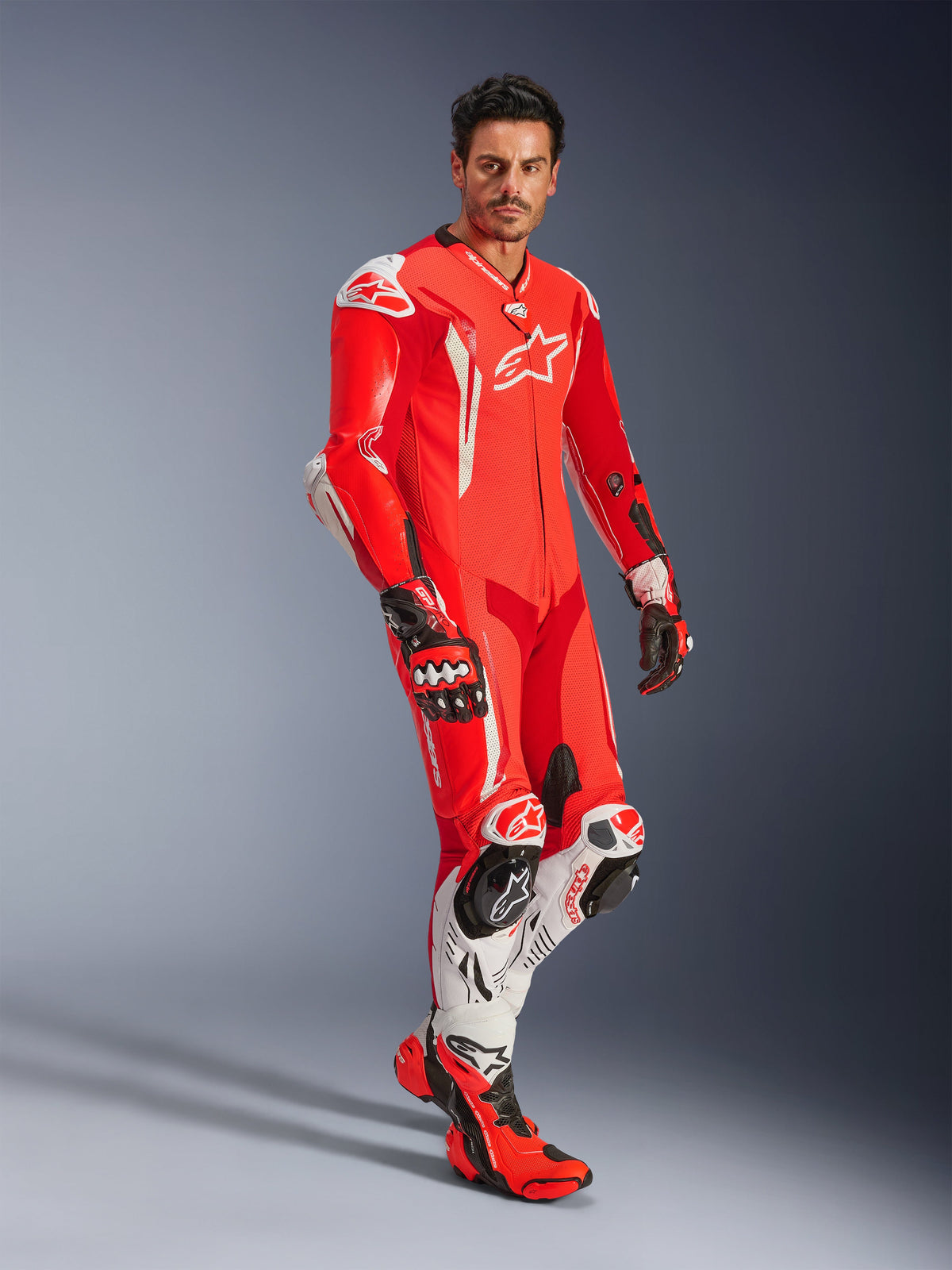 Homme portant une combinaison en cuir une pièce Alpinestars GP Tech V5, rouge fluo, rouge vif et blanc, coordonnée avec des gants et des bottes de course, équipement de moto professionnel doté d'une bosse aérodynamique et de protections intégrées.