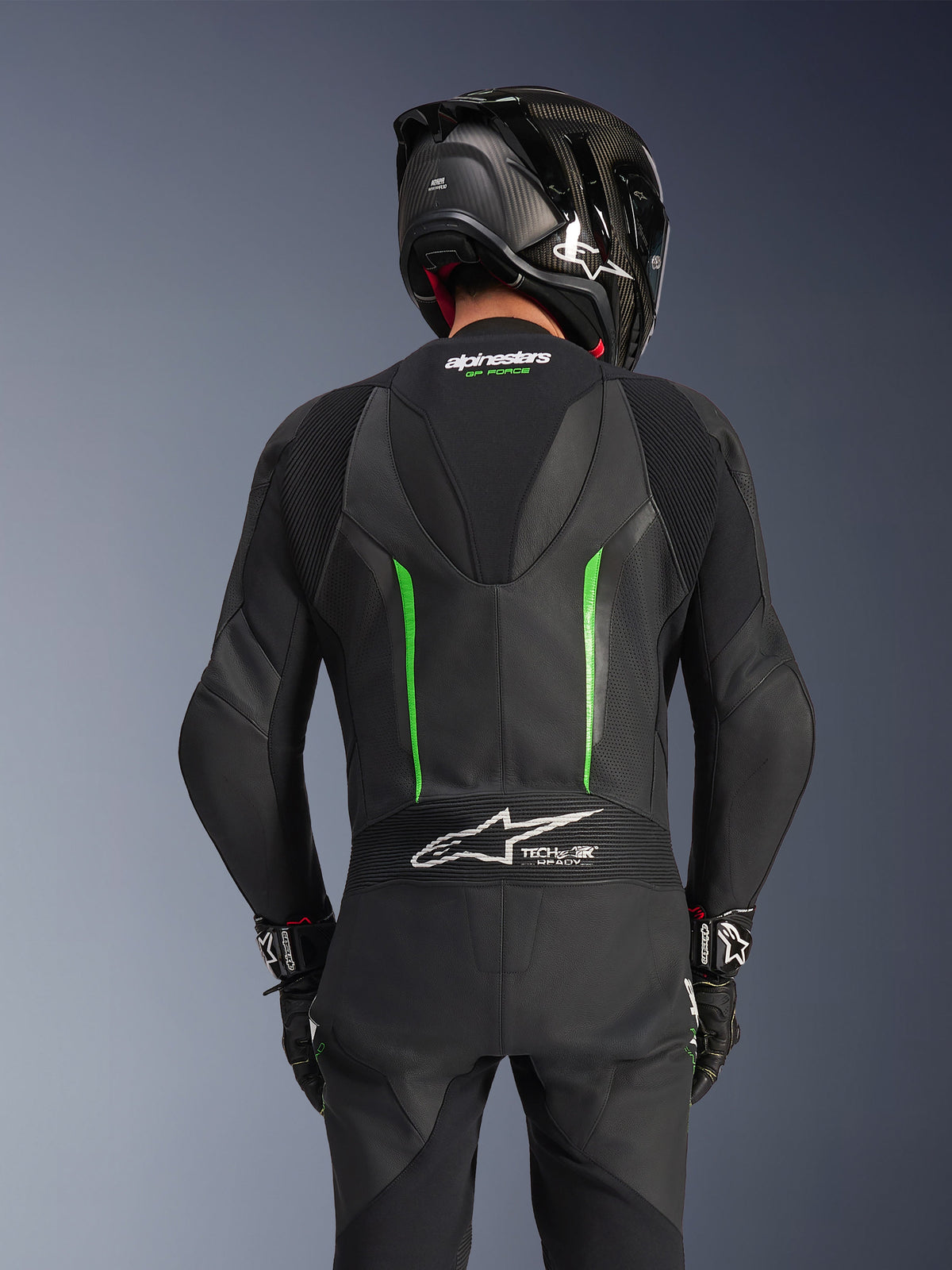 Rear view of a rider wearing the Alpinestars GP Force V2 Leather Suit in Black Green, featuring an aerodynamic speed hump, Tech-Air Ready branding on the lower back accordion panel, and perforated leather construction, paired with a black carbon fiber racing helmet.