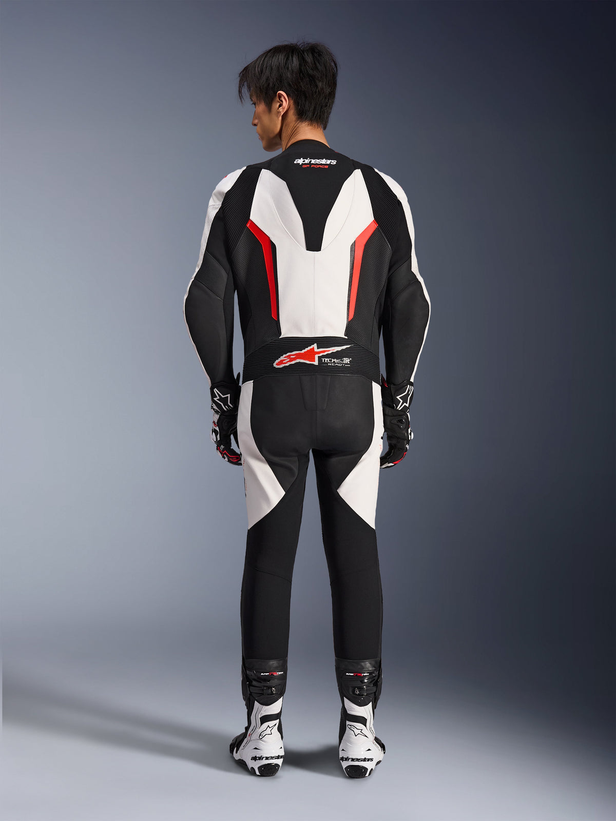 Combinaison en cuir Alpinestars GP Force V2 1 pièce, combinaison de course moto, blanc, noir et rouge fluo, vue arrière du mannequin portant la combinaison avec des bottes et des gants de course, dotée d'une bosse aérodynamique, de panneaux extensibles en accordéon et du logo Tech-Air Ready