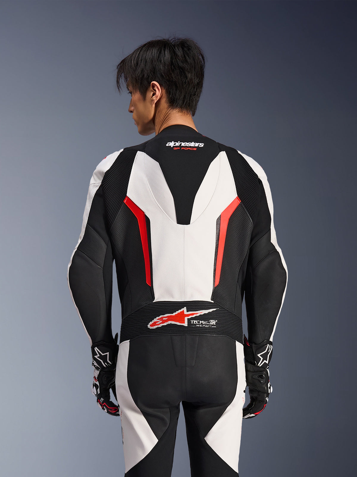 Combinaison en cuir Alpinestars GP Force V2 1 pièce, blanc, noir et rouge fluo, vue de dos sur un modèle masculin, avec bosse aérodynamique, panneaux en cuir perforé et logo Tech-Air Ready dans le bas du dos, équipement de course professionnel avec gants noirs et blancs