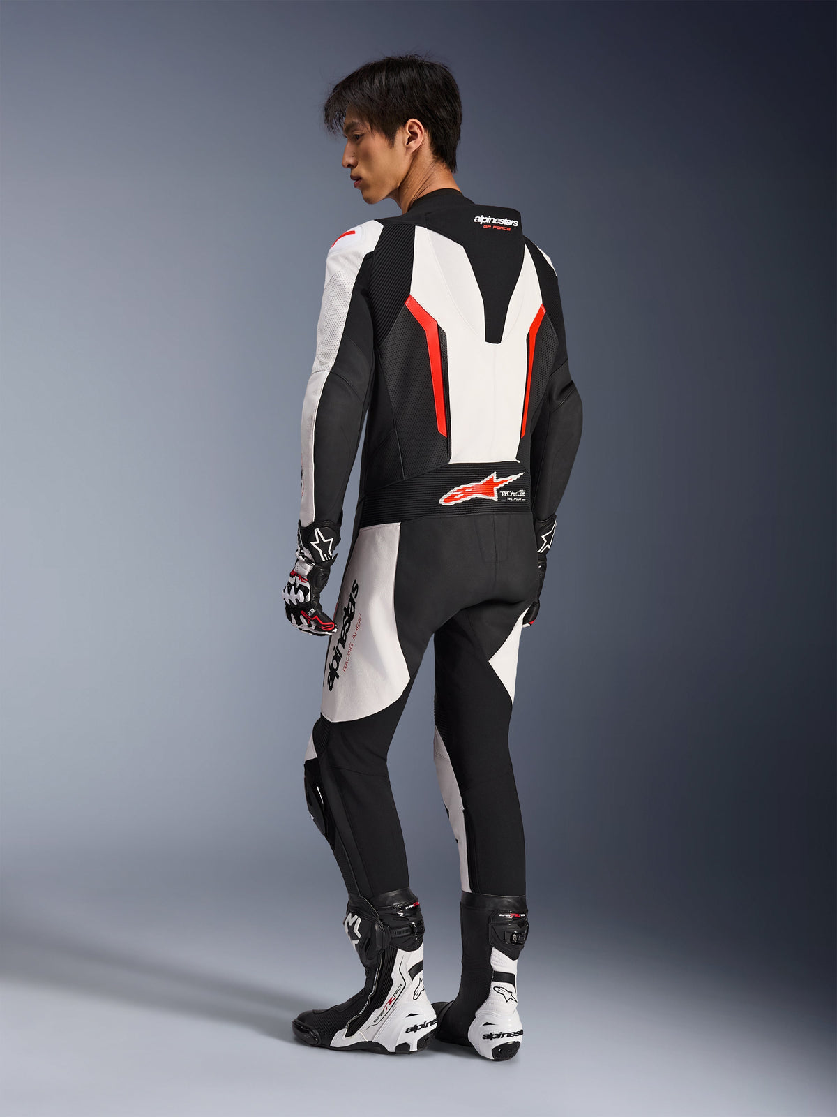 Pilote de moto portant la combinaison en cuir Alpinestars GP Force V2 - 1 pièce en blanc, noir et rouge fluo, vue arrière latérale montrant la bosse aérodynamique et les panneaux en cuir perforé, associée à des bottes et des gants de course assortis, conçue pour la piste et la performance.