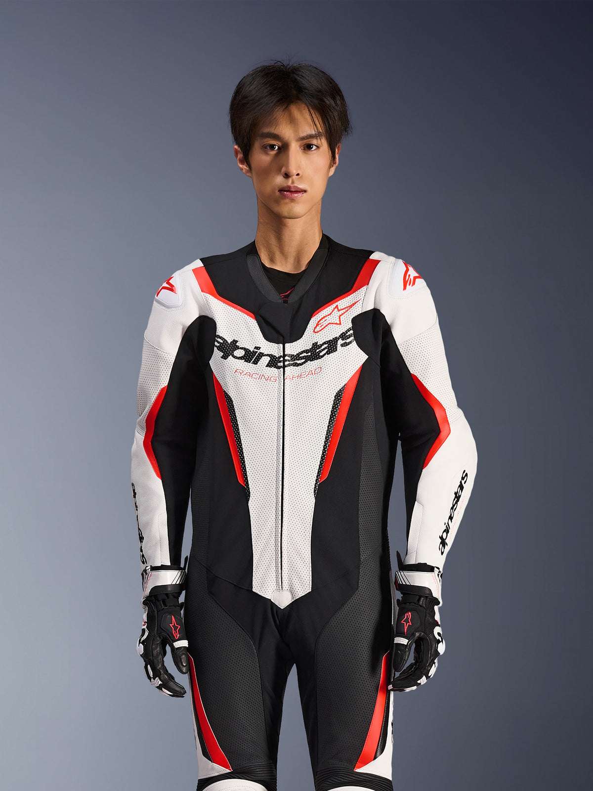 Combinaison en cuir Alpinestars GP Force V2 - 1 pièce, blanc, noir et rouge fluo, portée par un mannequin homme, avec panneaux en cuir perforé et logo Alpinestars sur la poitrine, assortie à des gants de course noirs et blancs, conçue pour la piste et la performance