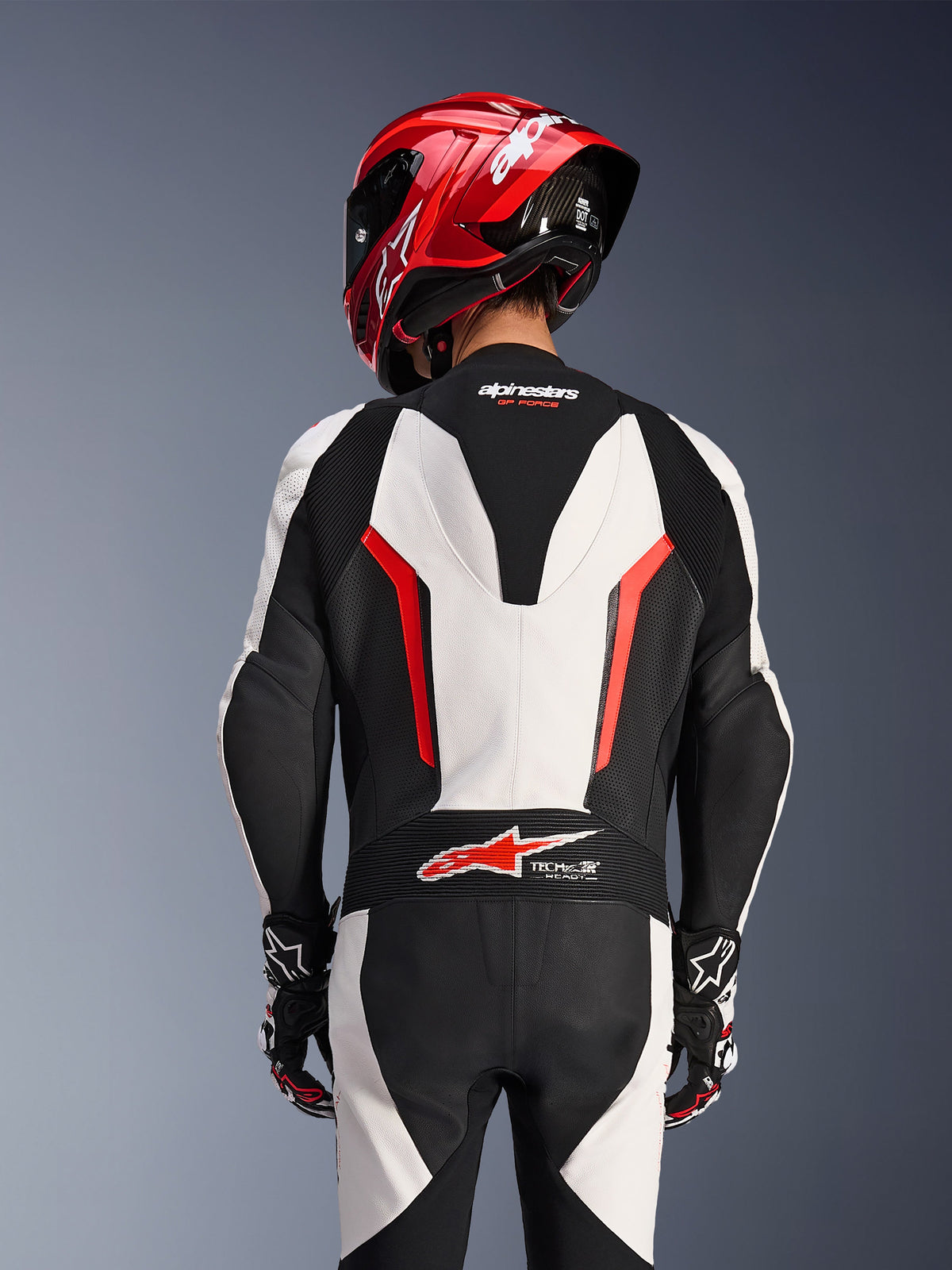 Vue arrière de la combinaison en cuir Alpinestars GP Force V2 1 pièce en blanc, noir et rouge fluo, portée avec un casque de course rouge, présentant le marquage Tech-Air Ready sur le bas du dos, des panneaux en cuir perforé et des zones d'extension en accordéon pour la performance en course.