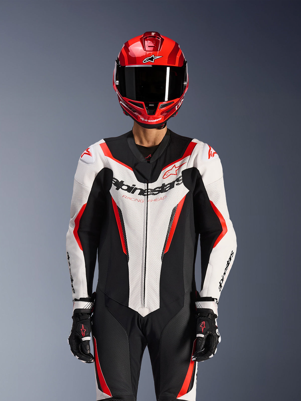 Combinaison en cuir Alpinestars GP Force V2 - 1 pièce, blanc, noir et rouge fluo, portée avec un casque intégral rouge et des gants de course noirs, panneaux en cuir perforé et logo Alpinestars sur la poitrine, équipement de performance professionnel pour la course moto