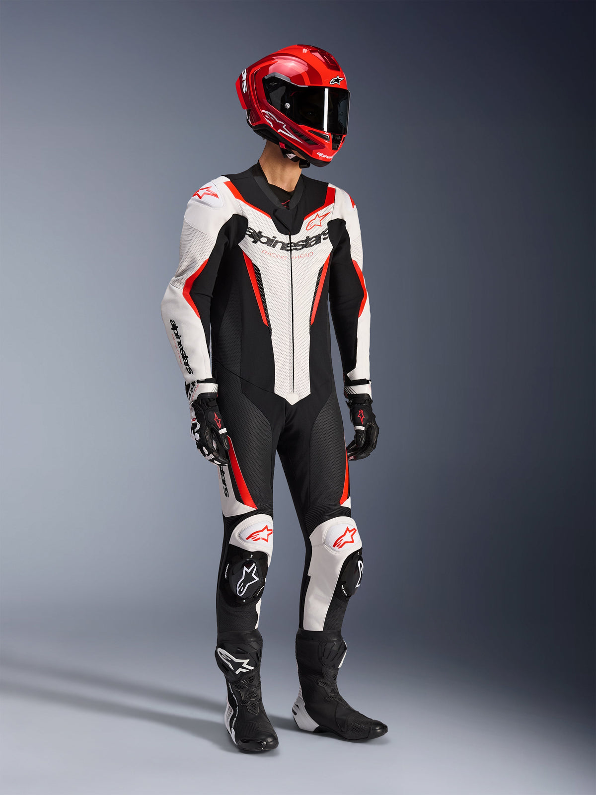 Combinaison en cuir Alpinestars GP Force V2 1 pièce en blanc, noir et rouge fluo, photo de studio en plein pied d'un pilote portant un équipement de course complet incluant un casque rouge, des gants noirs et des bottes noires, dotée d'une construction aérodynamique en cuir bovin avec marquage distinctif et sliders de genoux intégrés.