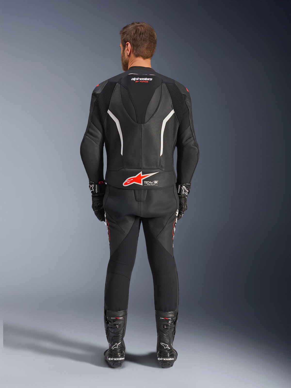 Vue de dos d'un pilote portant la combinaison en cuir 2 pièces Alpinestars GP Force V2, noire avec accents blancs et rouges, présentant des logos Alpinestars et Tech-Air Ready visibles au dos, accompagnée de gants et de bottes de course noirs assortis.