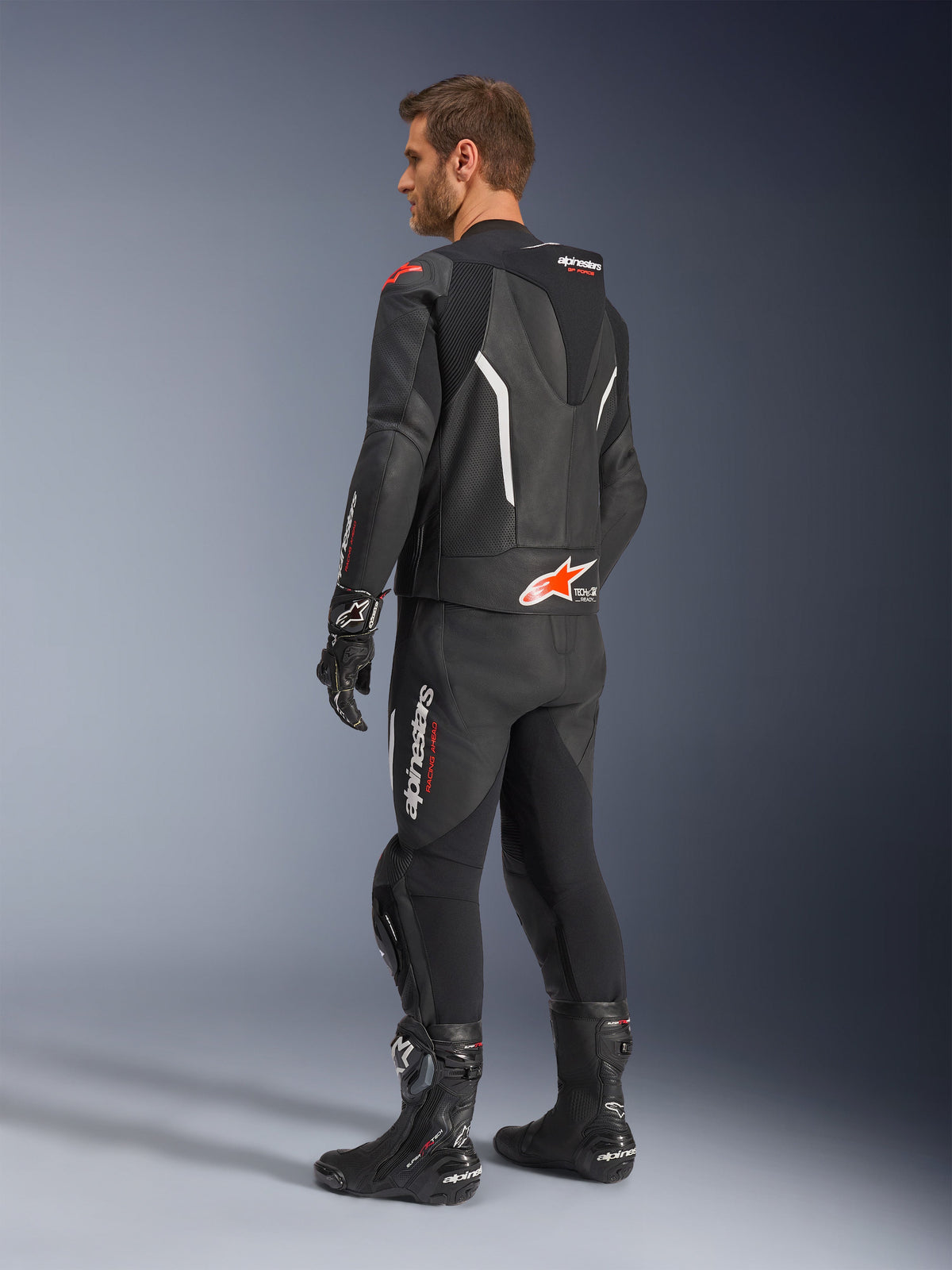 Combinaison en cuir 2 pièces Alpinestars GP Force V2, noire avec accents blancs et rouge fluo, vue arrière de trois-quarts sur modèle, dotée d'une bosse aérodynamique, de panneaux en cuir perforé et d'un marquage Alpinestars distinctif, portée avec des bottes et des gants de course noirs.