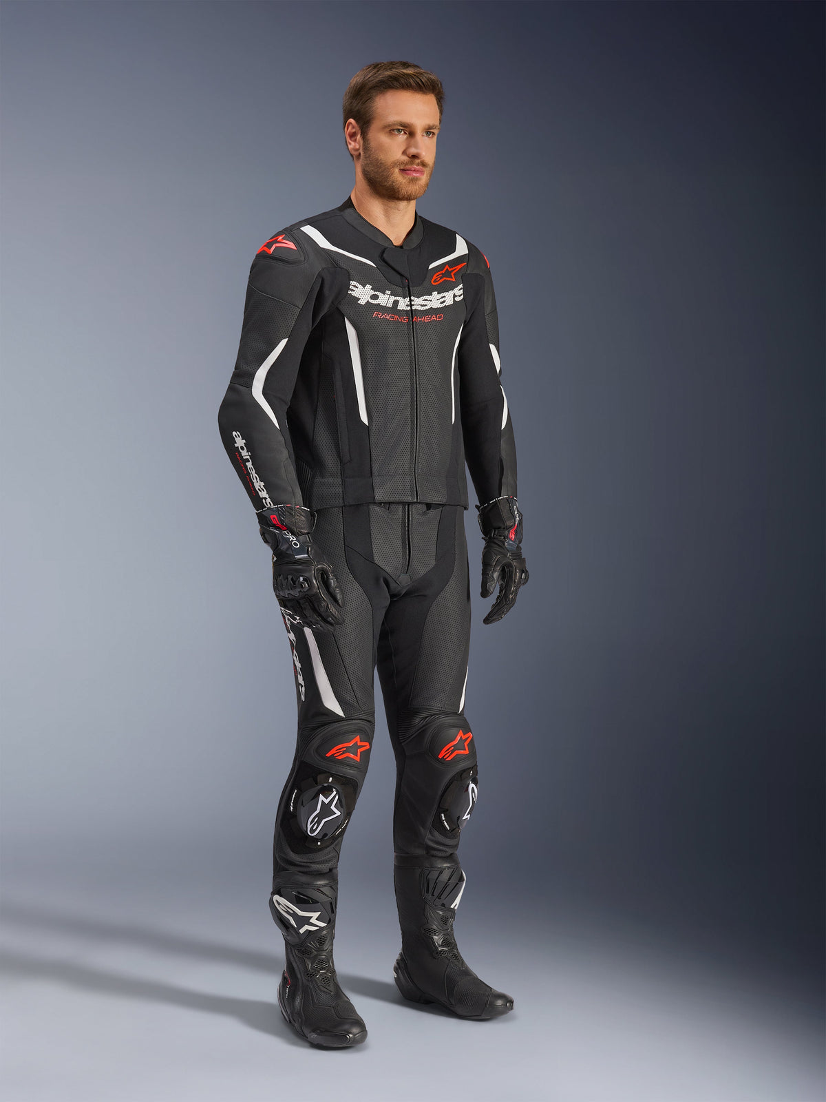 Combinaison en cuir 2 pièces Alpinestars GP Force V2, noir, blanc et rouge fluo, portée par un mannequin avec des bottes et gants de course noirs, avec panneaux en cuir perforé, accents aérodynamiques blancs et logos rouge fluo sur les épaules et les genoux, équipement de course moto professionnel
