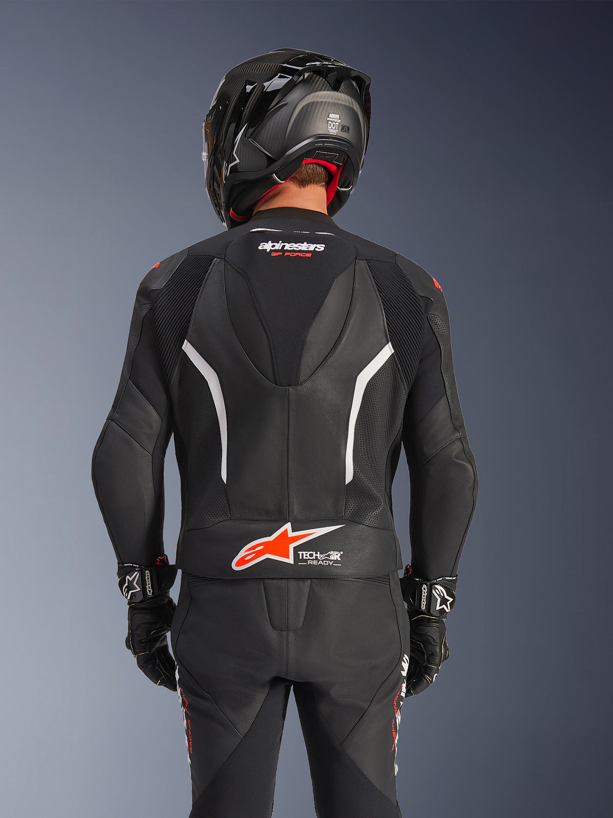 Vue arrière de la combinaison en cuir 2 pièces Alpinestars GP Force V2 en noir, avec cuir perforé, accents blancs et logo Alpinestars rouge sur le bas du dos, portée avec un casque de moto noir et des gants de course.