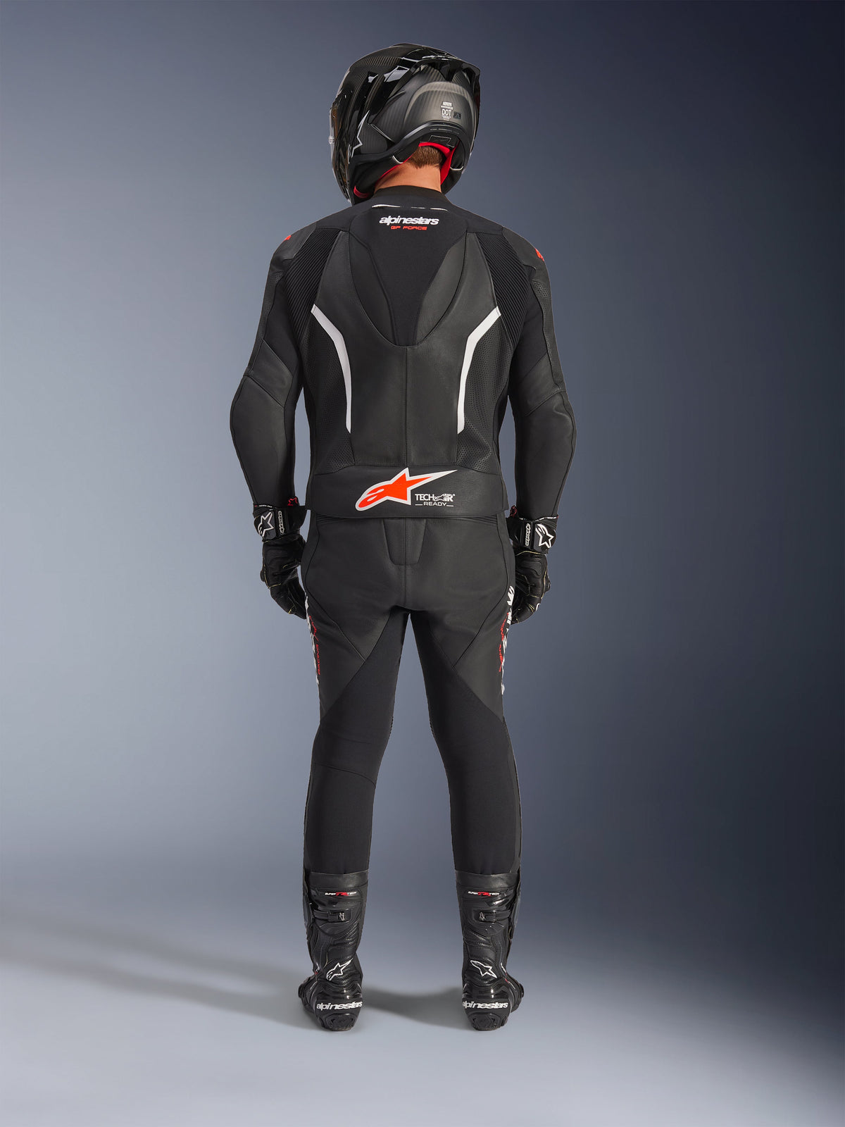 Motard vu de dos portant la combinaison en cuir 2 pièces Alpinestars GP Force V2, noire avec des accents blanc et rouge fluo, dotée d'une bosse aérodynamique et du logo Tech-Air Ready, avec casque de course, gants et bottes noirs.