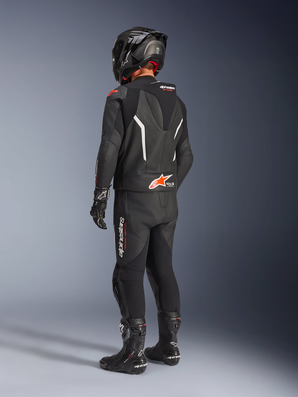 Combinaison en cuir 2 pièces Alpinestars GP Force V2, combinaison de moto en cuir noir vue de dos, portée avec un casque, des gants et des bottes noirs assortis, détails blancs et rouges, bosse aérodynamique avec logo Alpinestars, équipement de course et de sport professionnel.