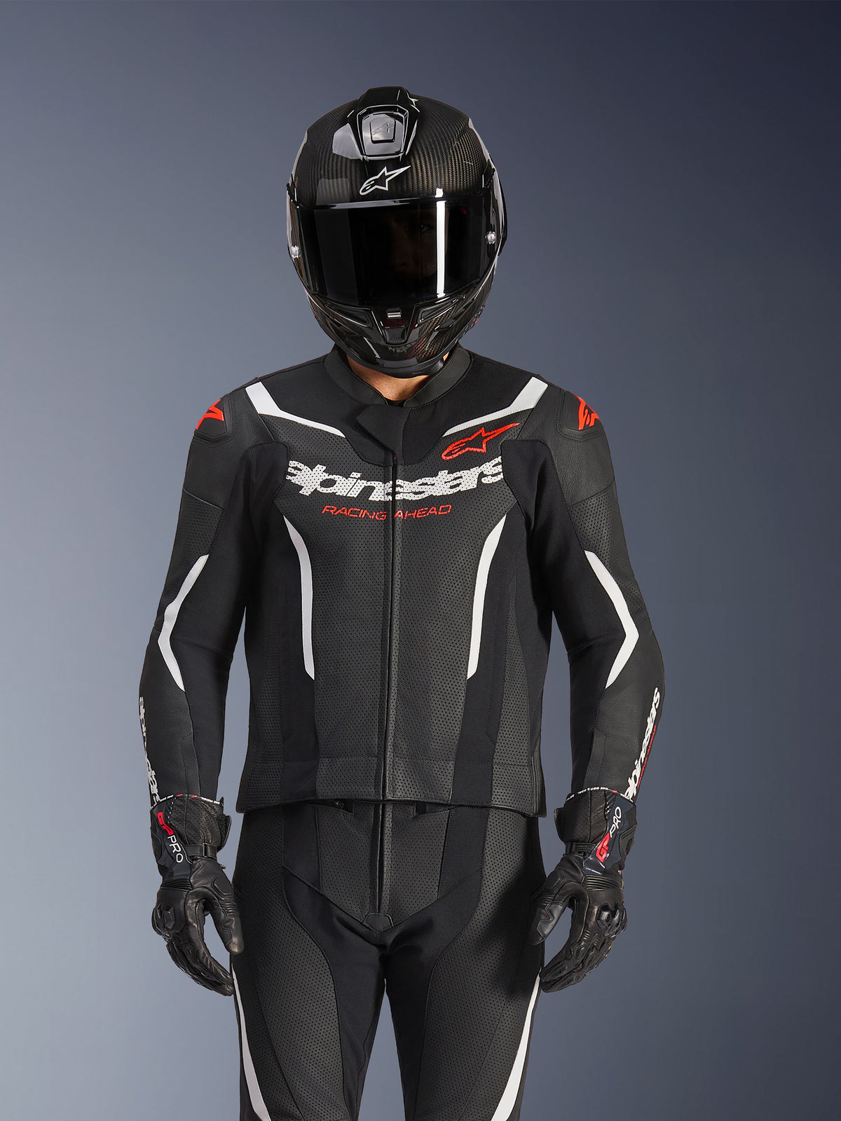 Combinaison en cuir 2 pièces Alpinestars GP Force V2, noir, blanc et rouge fluo, portée par un pilote avec casque intégral en fibre de carbone et gants de course noirs, avec panneaux en cuir perforé et logo Alpinestars sur la poitrine.