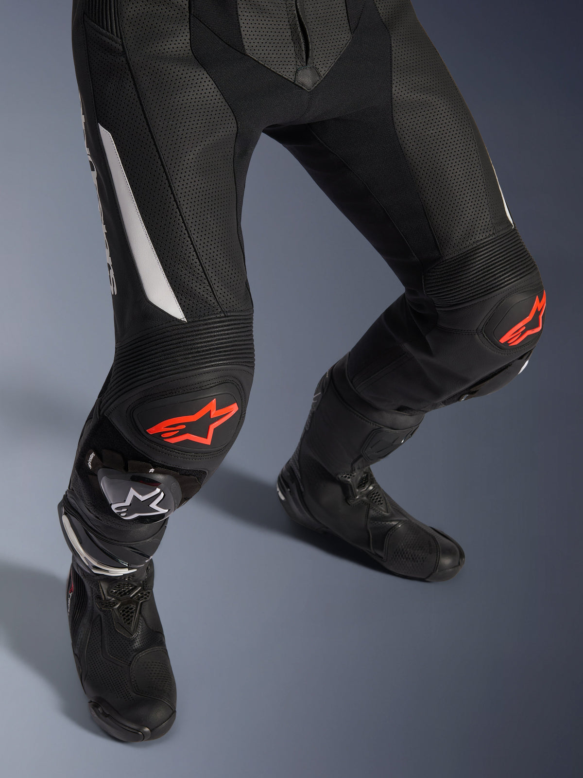 Combinaison en cuir Alpinestars GP Force V2 2 pièces, pantalon de course moto noir avec accents rouges et blancs, doté de cuir perforé, panneaux stretch en accordéon au-dessus des genoux avec logos d'étoiles rouges, porté avec des bottes de moto noires, équipement de course haute performance