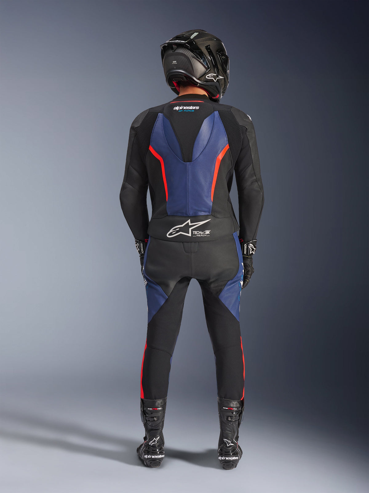 Alpinestars GP Force V2 Leather Suit 2-Piece, Black Blue Red Fluo, full rear view of a rider in complete racing gear, featuring blue leather panels on the back and legs with fluorescent red accents, Tech-Air® Ready logo on the lower back, paired with a carbon fiber helmet, black gloves, and racing boots.