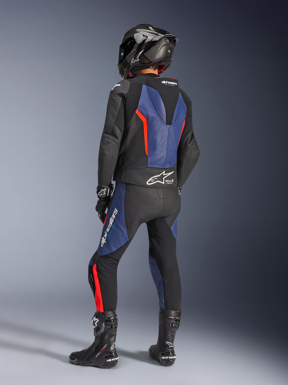 Alpinestars GP Force V2 Leather Suit 2-Piece, Black Blue Red Fluo, rear view of rider in professional racing gear, featuring an integrated speed hump and Tech-Air Ready branding, blue and red fluo accents on black leather, worn with a carbon fiber helmet and black boots, high-performance motorcycle apparel