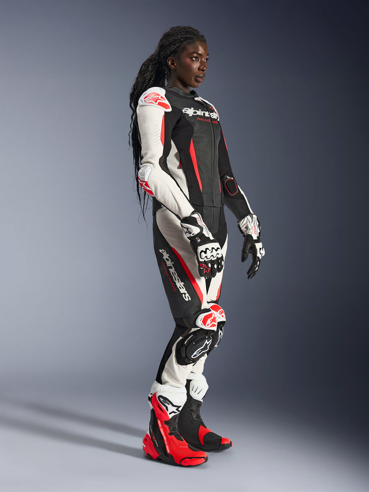 Femme pilote portant la combinaison en cuir 2 pièces Alpinestars Stella GP-R7 en noir, blanc et rouge fluo, avec gants et bottes de course assortis, dotée de panneaux en cuir perforé et de protections aux épaules, coudes et genoux, dans un studio.