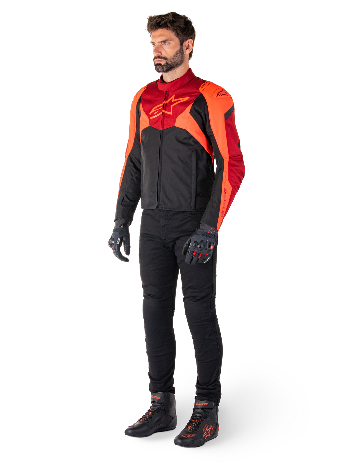Motard portant la veste imperméable Alpinestars T-Jaws V4 en Noir Rouge Fluo Rouge Foncé, accompagnée d'un pantalon de moto, de gants et de bottes de pilotage noirs, dotée d'un design textile à coupe sport avec un marquage imposant sur la poitrine et une silhouette agressive.