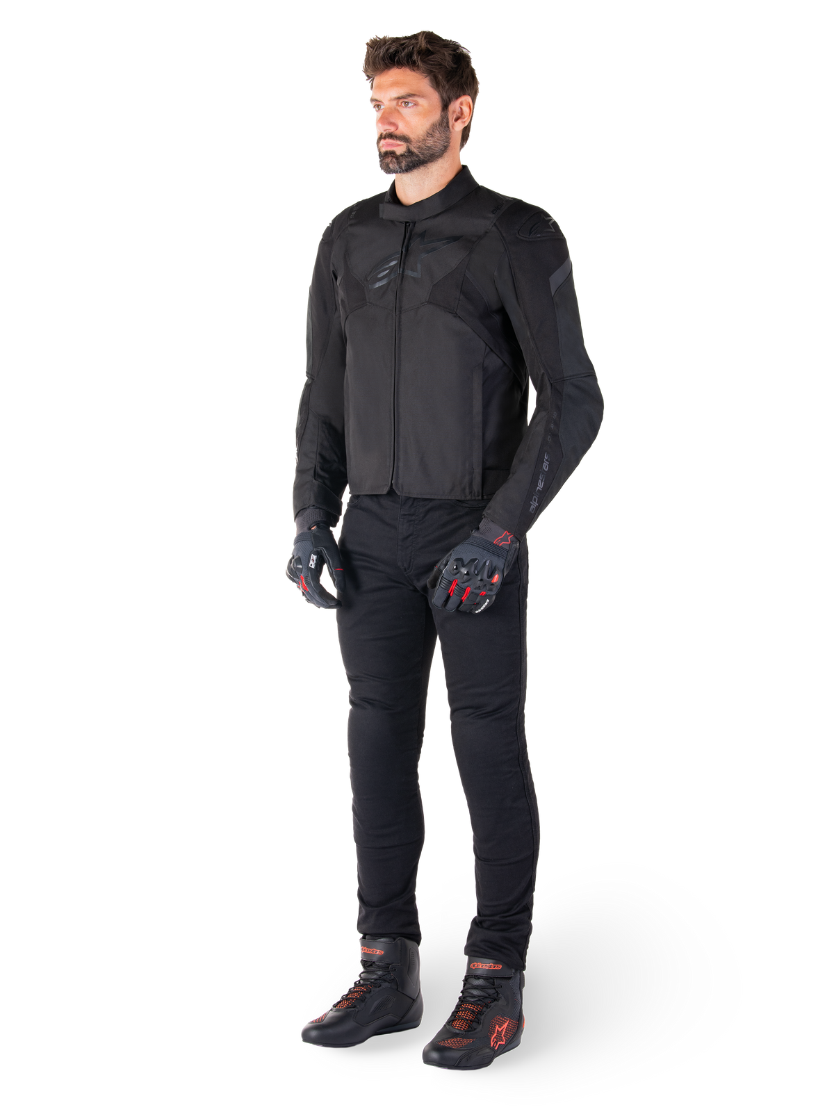 Modèle masculin portant la veste imperméable Alpinestars T-Jaws V4 en noir, associée à un pantalon de moto noir, des gants noirs avec des accents rouges et des chaussures de moto noires, présentant une coupe sport élégante et un marquage discret, isolé sur fond blanc.