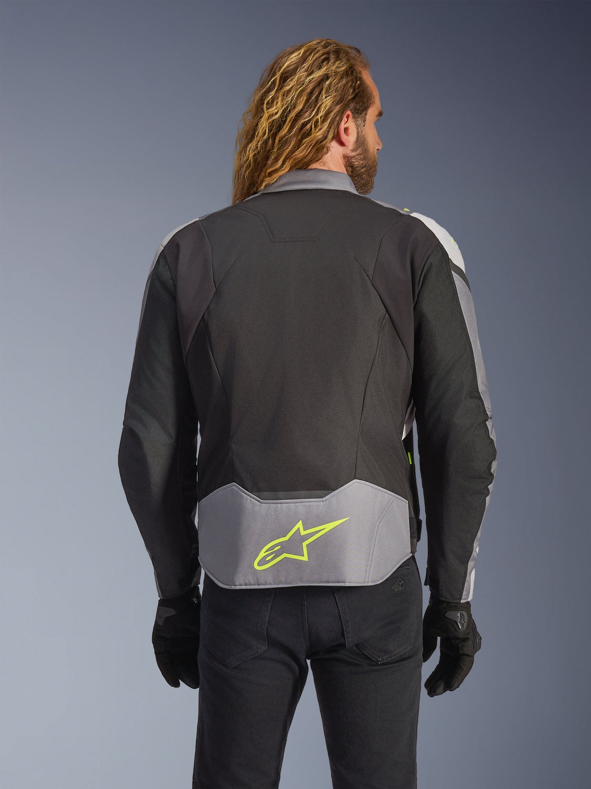 Veste imperméable Alpinestars T-Jaws V4, Noir Gris Glacé Gris Foncé, vue arrière sur modèle, dotée d'un empiècement gris clair au bas du dos avec un logo étoile jaune fluo distinctif, construction textile multi-panneaux et silhouette sportive, portée avec des gants noirs et un pantalon sombre.