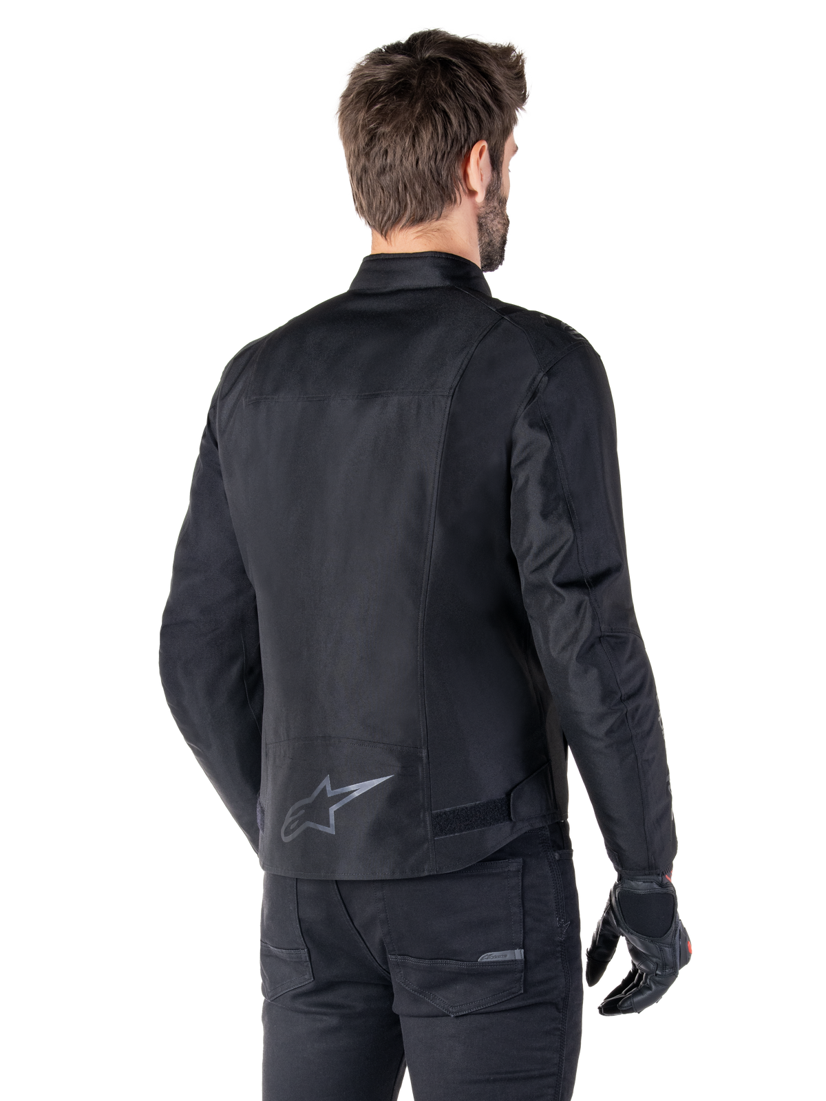 Veste imperméable Alpinestars T-SPS V2, noire, vue arrière sur un mannequin homme, avec un logo étoile réfléchissant dans le bas du dos, un profil arrière allongé et des sangles de réglage à la taille, portée avec un pantalon de moto et des gants noirs