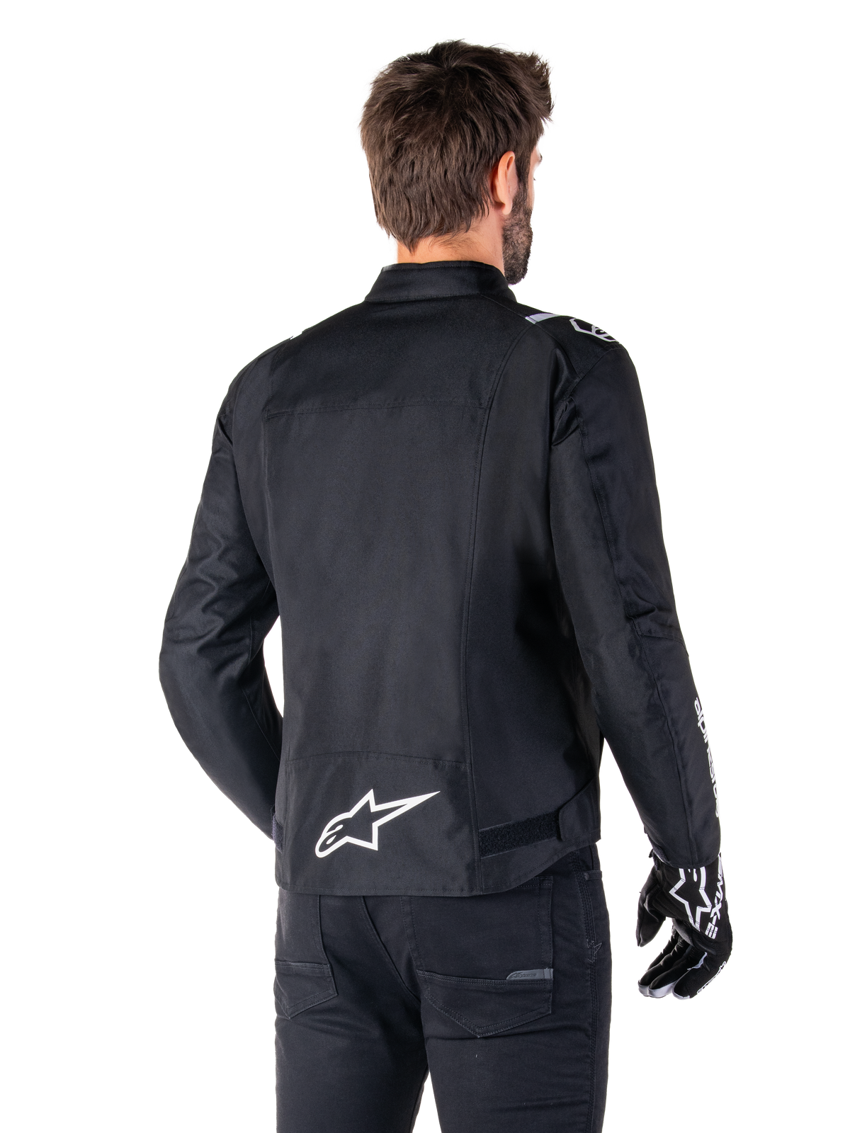Veste imperméable Alpinestars T-SPS V2, noir et blanc, vue arrière d'un motard portant la veste, logo Alpinestars blanc dans le bas du dos, construction textile à coupe sport, portée avec un pantalon et des gants de moto noirs