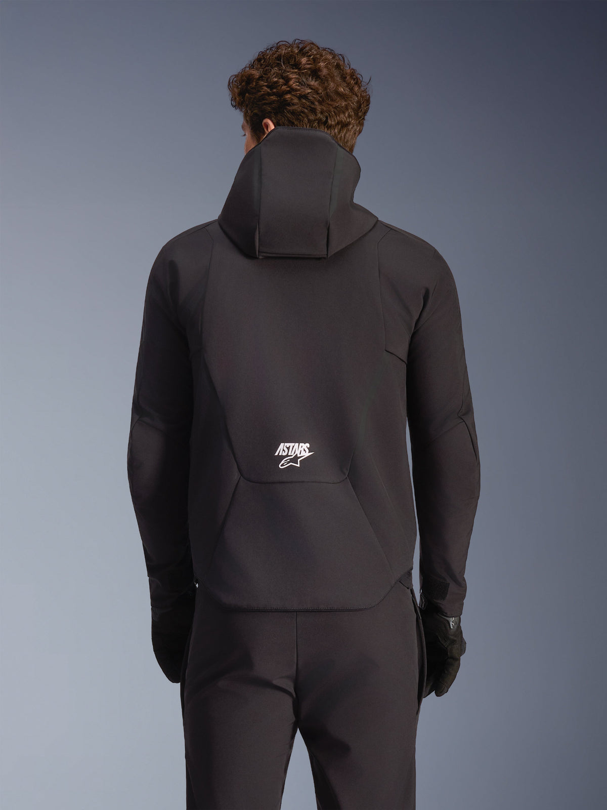 Alpinestars Aeroshell Waterproof Jacket, black road motorcycle softshell, rear view on model with hood and white logo.