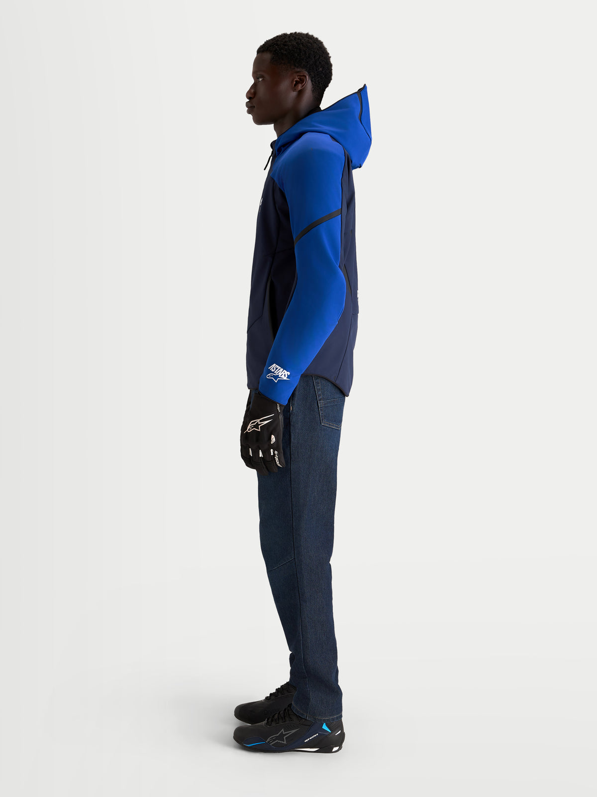 Veste imperméable Aeroshell d'Alpinestars, veste de moto pour homme bleu nuit et bleu vif avec capuche et construction en softshell extensible, portée par un modèle en vue de profil