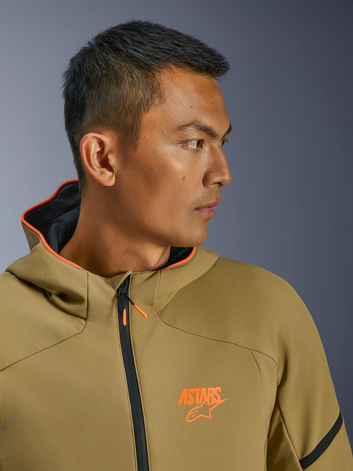 Alpinestars Aeroshell Waterproof Jacket, Dark Khaki and Orange Fluo, close-up side profile, featuring orange ASTARS logo on the chest and orange-lined hood, modern crossover softshell hoodie for motorcycle riding