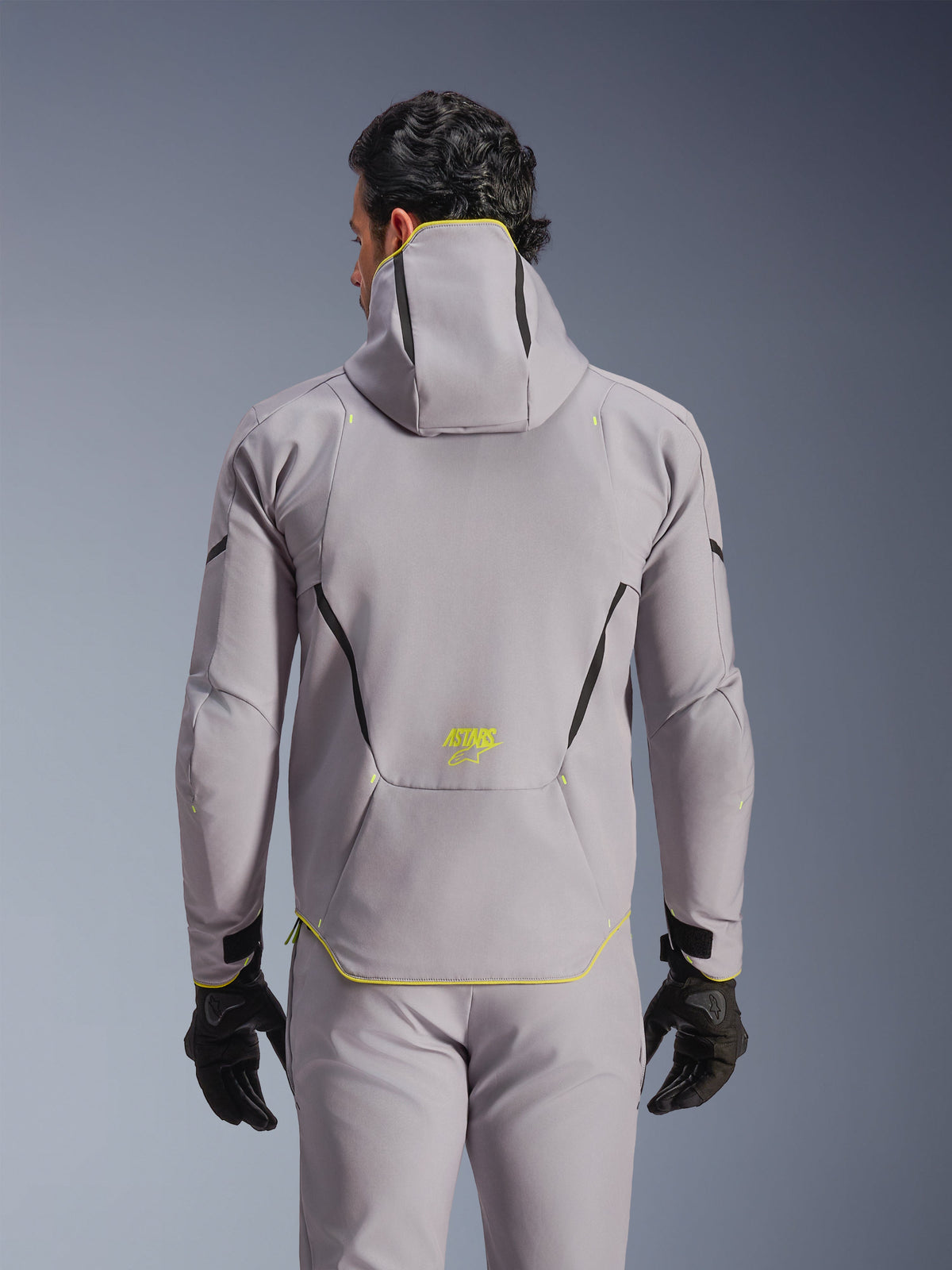 Veste Alpinestars Aeroshell WP, gris moyen avec accents jaune fluo, vue de dos montrant la capuche intégrée et des lignes de contraste noires, logo ASTARS jaune en bas du dos, portée avec un pantalon gris assorti et des gants noirs, veste de moto softshell sportive