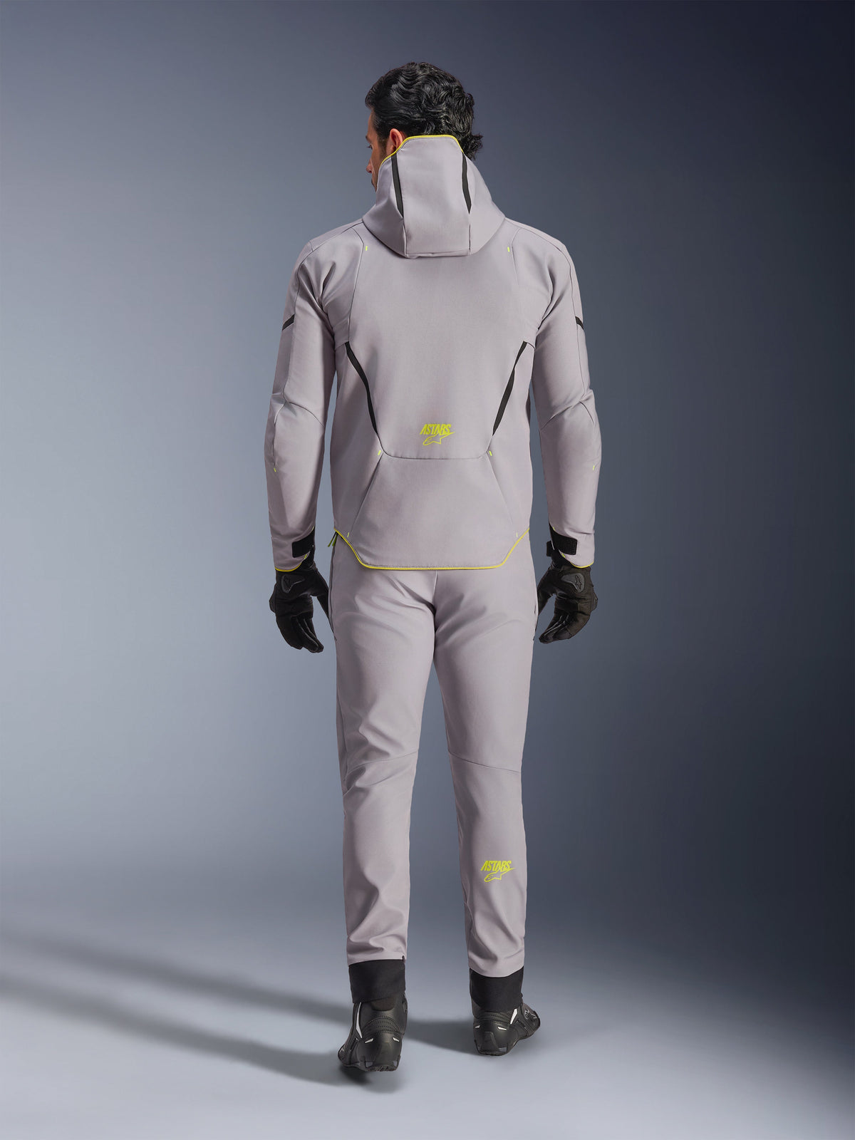 Veste imperméable Alpinestars Aeroshell en Mid Gray Yellow Fluo, vue de dos complète, portée avec un pantalon gris assorti, des gants noirs et des chaussures de moto noires. Modèle softshell à capuche avec marquage et accents 'ASTARS' jaune fluo, conçu pour la performance routière moderne à moto.