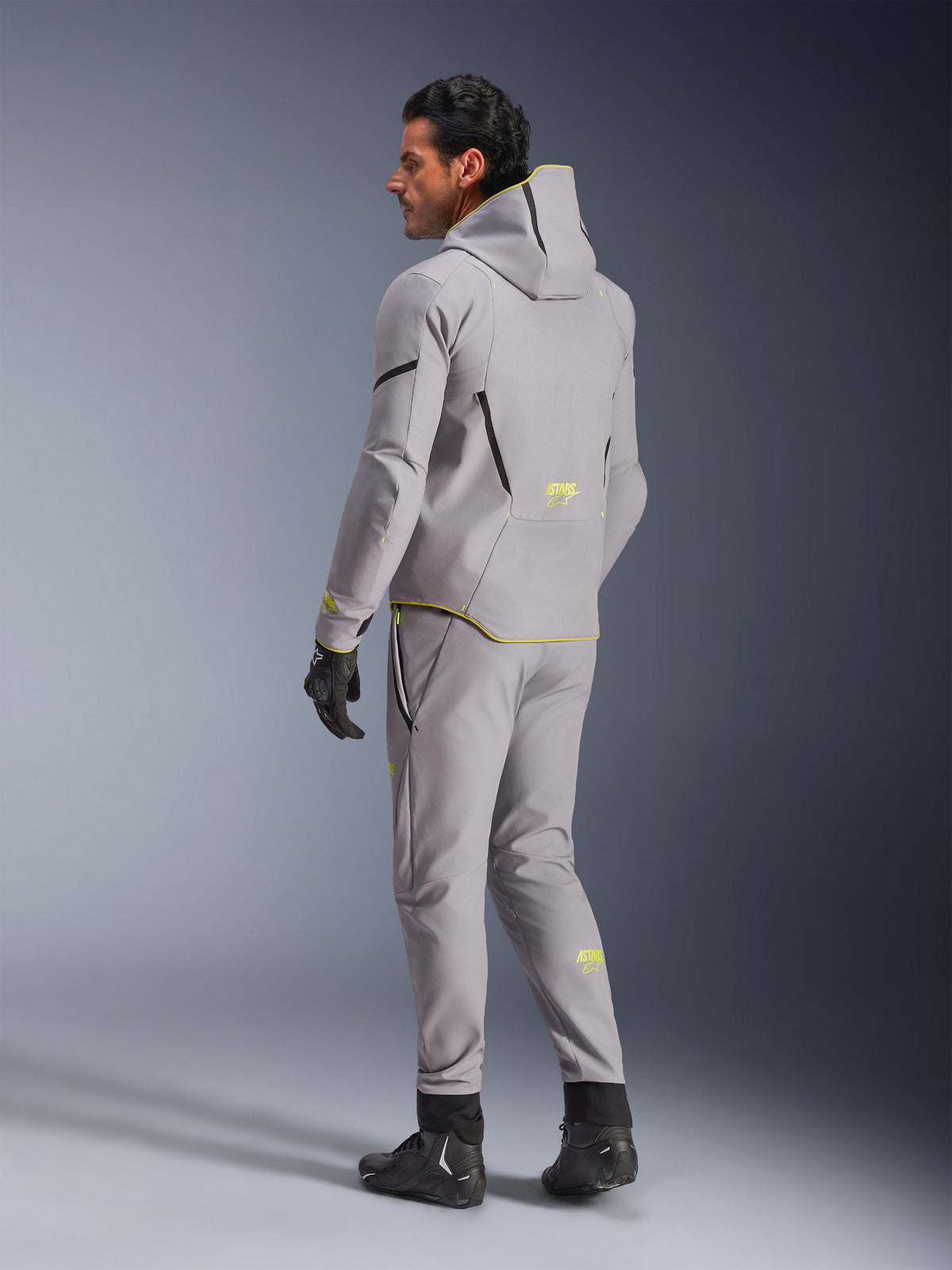 Veste imperméable Alpinestars Aeroshell, gris moyen jaune fluo, portée avec un pantalon gris assorti, présente un design à capuche avec des accents et logos jaunes, vue de dos, associée à des gants et des bottes de moto noirs, vêtement de moto moderne