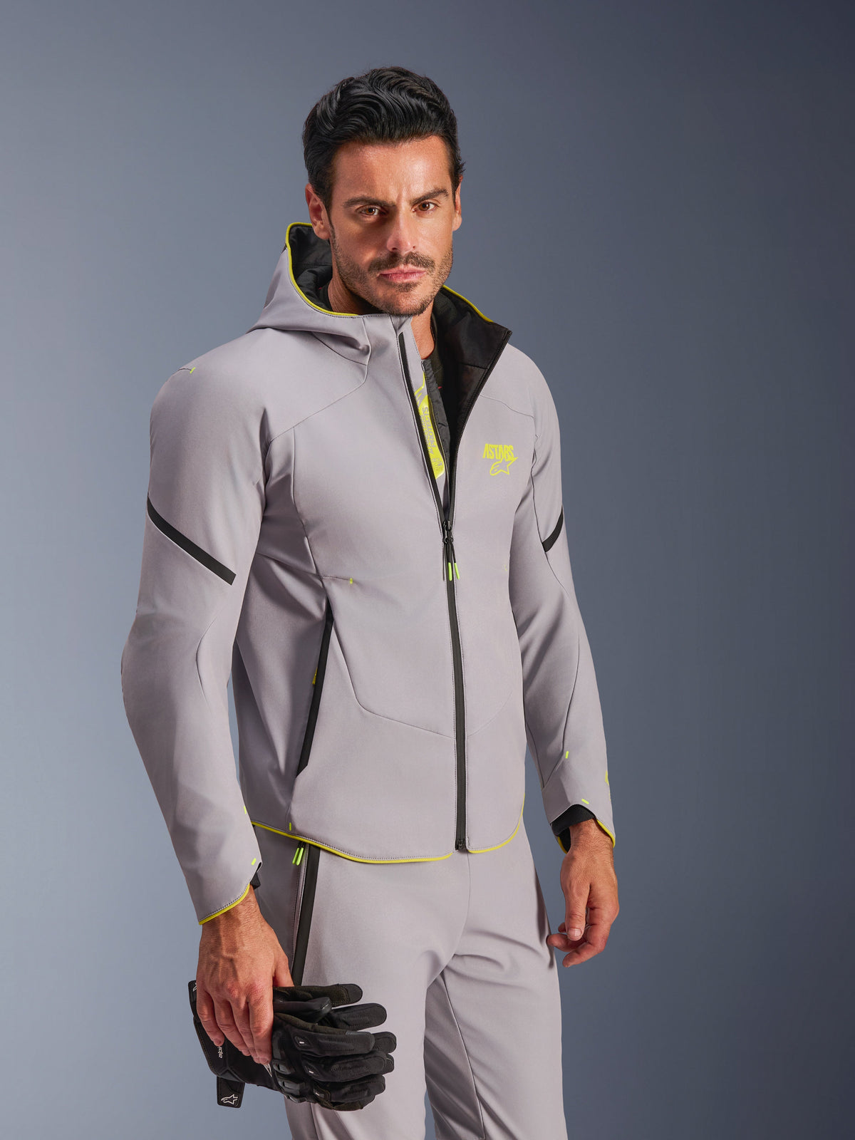 Veste imperméable Alpinestars Aeroshell en gris moyen et jaune fluo, portée par un homme avec un pantalon gris assorti et des gants de moto noirs, conception softshell à capuche avec accents jaune fluo et logo sur la poitrine, équipement de moto sportif moderne.