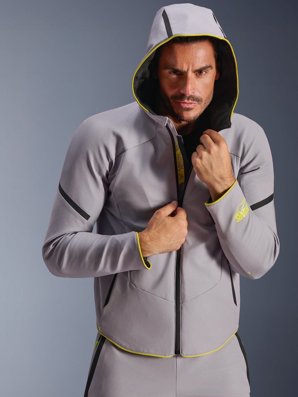 Veste imperméable Alpinestars Aeroshell, gris moyen et jaune fluo, veste softshell de sport avec capuche, portée par un mannequin homme avec la capuche relevée, dotée de fermetures éclair étanches noires et de touches jaune fluo sur les poignets et l'ourlet, design moderne inspiré par les sports mécaniques