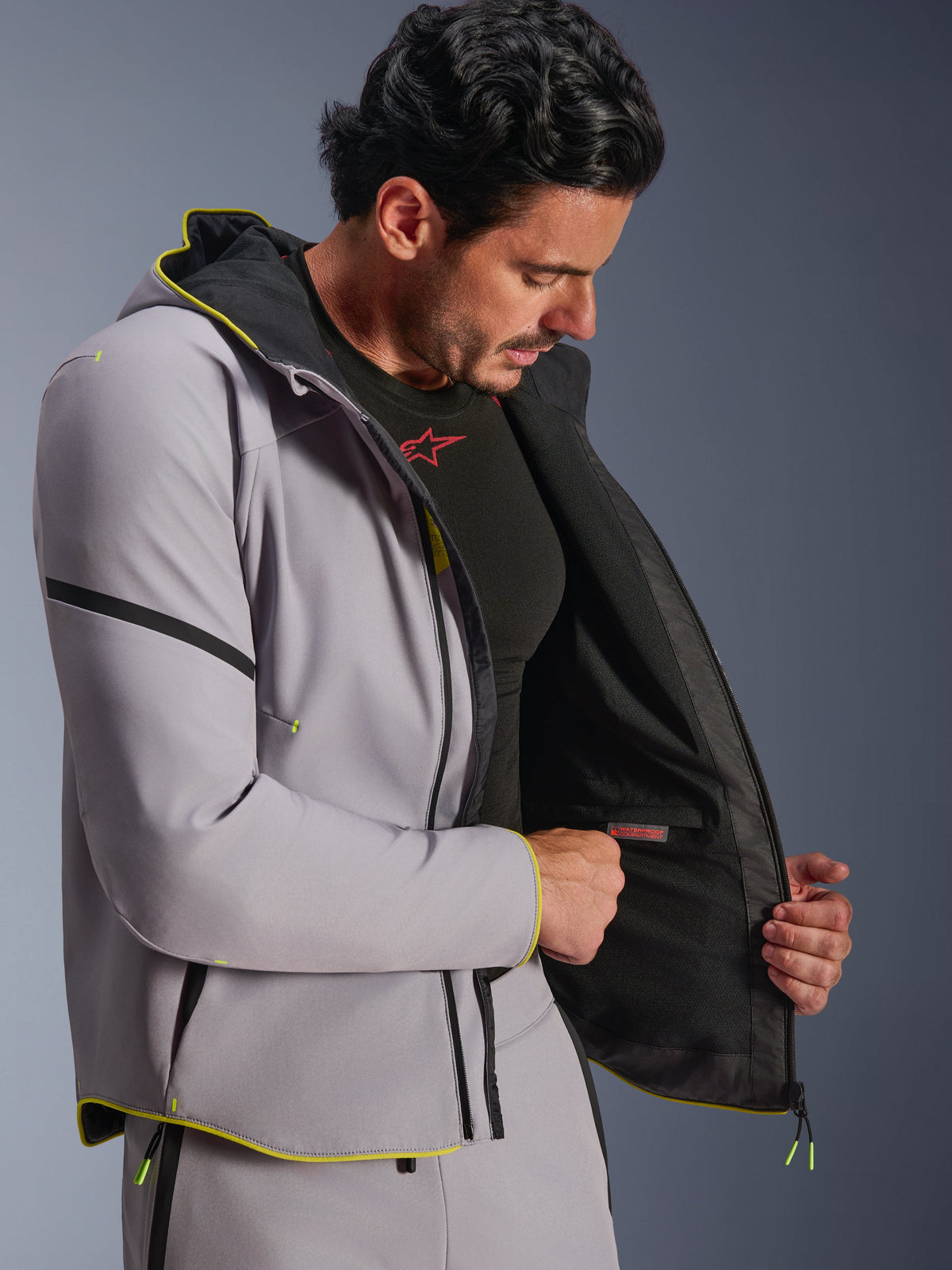Veste imperméable Alpinestars Aeroshell, gris moyen et jaune fluo, portée par un mannequin homme montrant le compartiment interne étanche et la doublure noire, hoodie de moto sport en softshell avec détails jaune fluo et coupe athlétique moderne.
