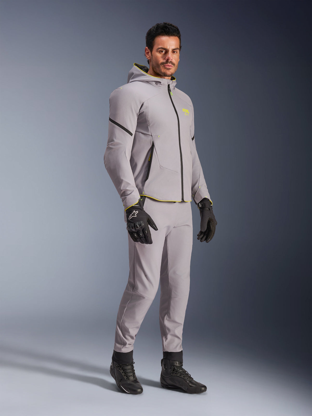 Veste imperméable Alpinestars Aeroshell, gris moyen et jaune fluo, portée par un mannequin avec un pantalon gris assorti, gants de moto noirs et bottes noires, équipement de moto moderne de style sweat à capuche pour une conduite décontractée et sportive