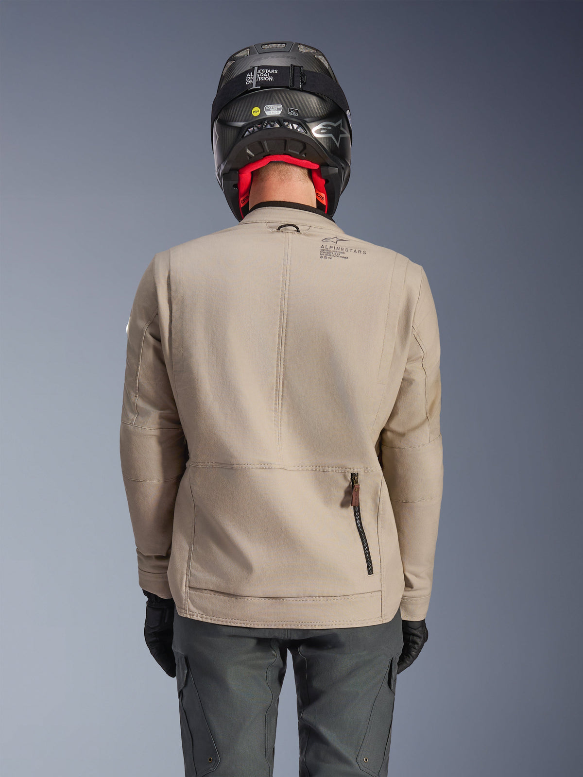 Veste Alpinestars Borrego Drystar®, veste de moto d'aventure, gris chaud, vue de dos montrant la fermeture éclair du compartiment arrière et le marquage Alpinestars, portée avec un casque noir et un pantalon de moto anthracite