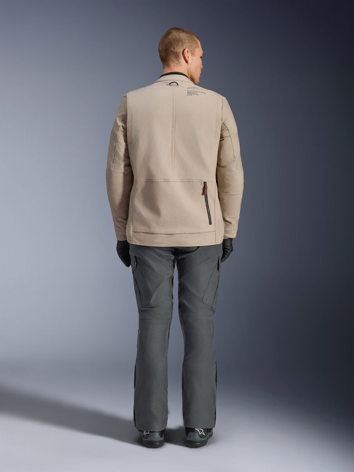 Veste Alpinestars Borrego Drystar®, vue arrière d'un mannequin homme portant la veste de moto textile Warm Gray, associée à un pantalon technique gris foncé, des gants noirs et des bottes, présentant le design robuste en toile cirée et des panneaux d'extension à l'arrière.