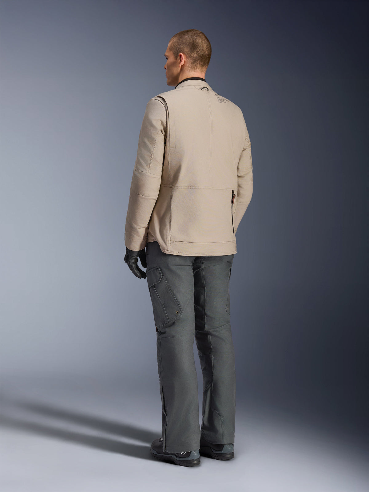 Veste Alpinestars Borrego Drystar® en coloris Warm Gray, vue arrière montrant la texture robuste en toile cirée, les panneaux d'expansion aux épaules et la grande poche arrière zippée, portée par un mannequin avec un pantalon d'aventure gris anthracite et des gants noirs.