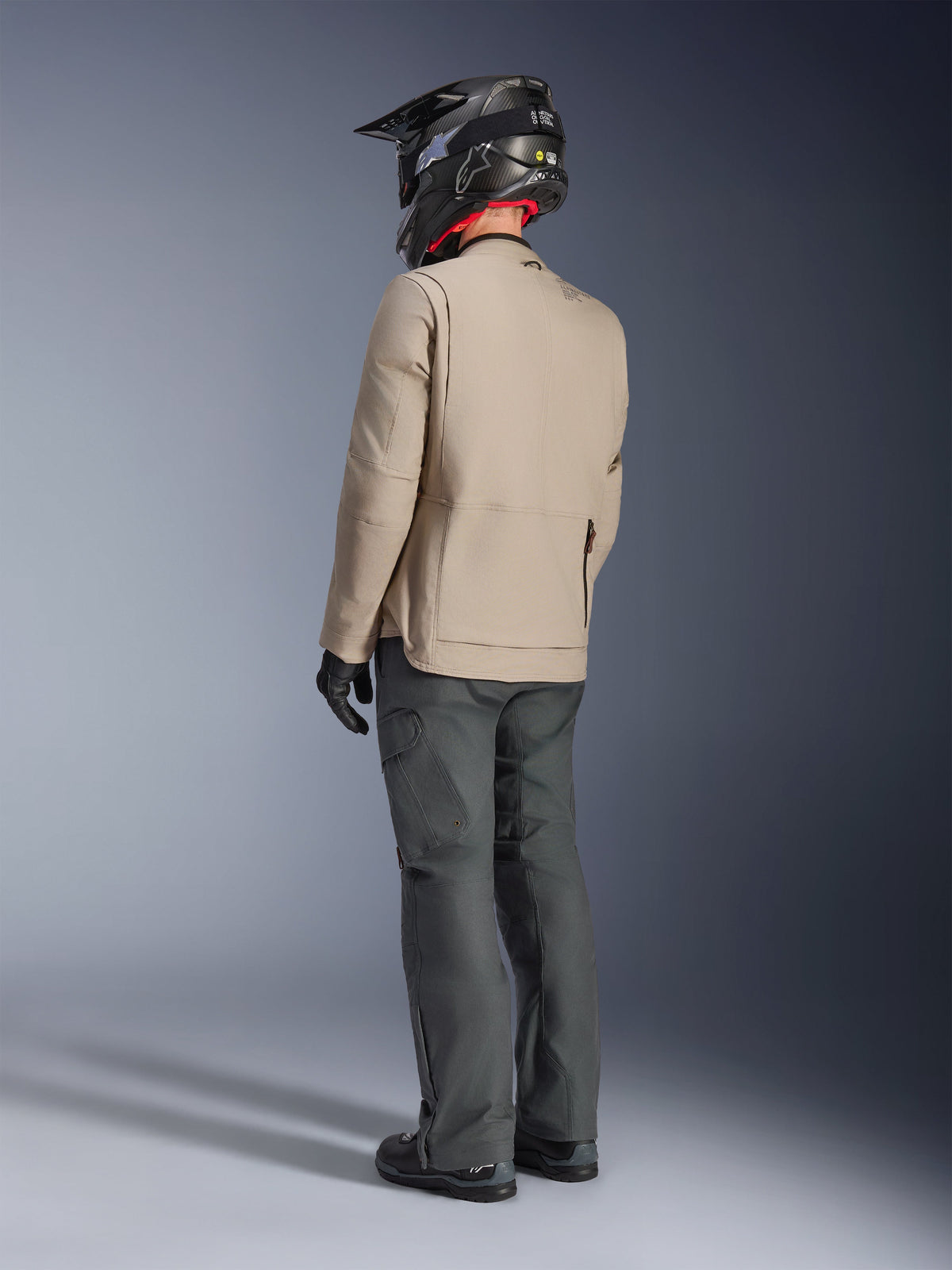 Veste Alpinestars Borrego Drystar®, veste de moto d'aventure gris chaud, vue de dos d'un pilote portant un pantalon gris foncé et un casque noir, présentant un aspect toile cirée robuste et un design fonctionnel.