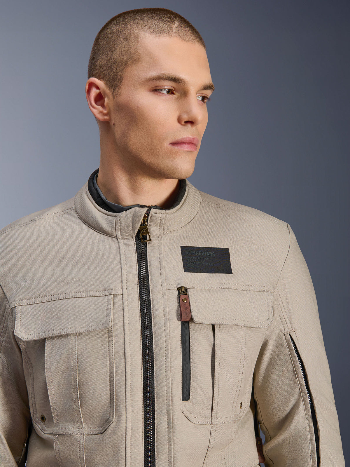 Modèle masculin portant la veste Alpinestars Borrego Drystar® en Warm Gray, veste de moto d'aventure avec un look classique en toile cirée, poches cargo fonctionnelles sur la poitrine et patch logo Alpinestars noir.