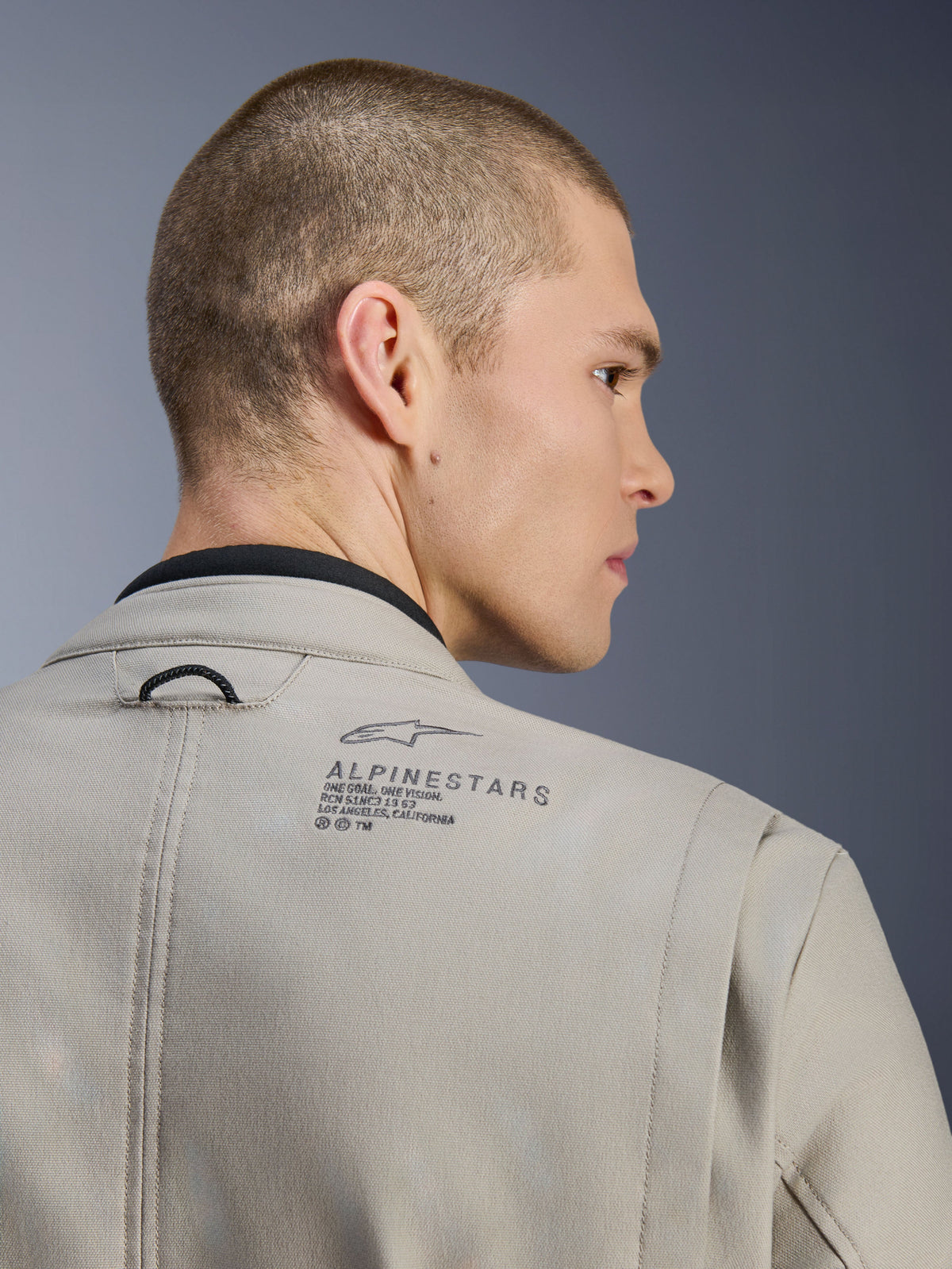 Veste Alpinestars Borrego Drystar® en Warm Gray, gros plan sur l'épaule arrière, matériau en toile cirée texturée, avec logo Alpinestars noir et texte héritage, portée par un mannequin, veste de moto d'aventure