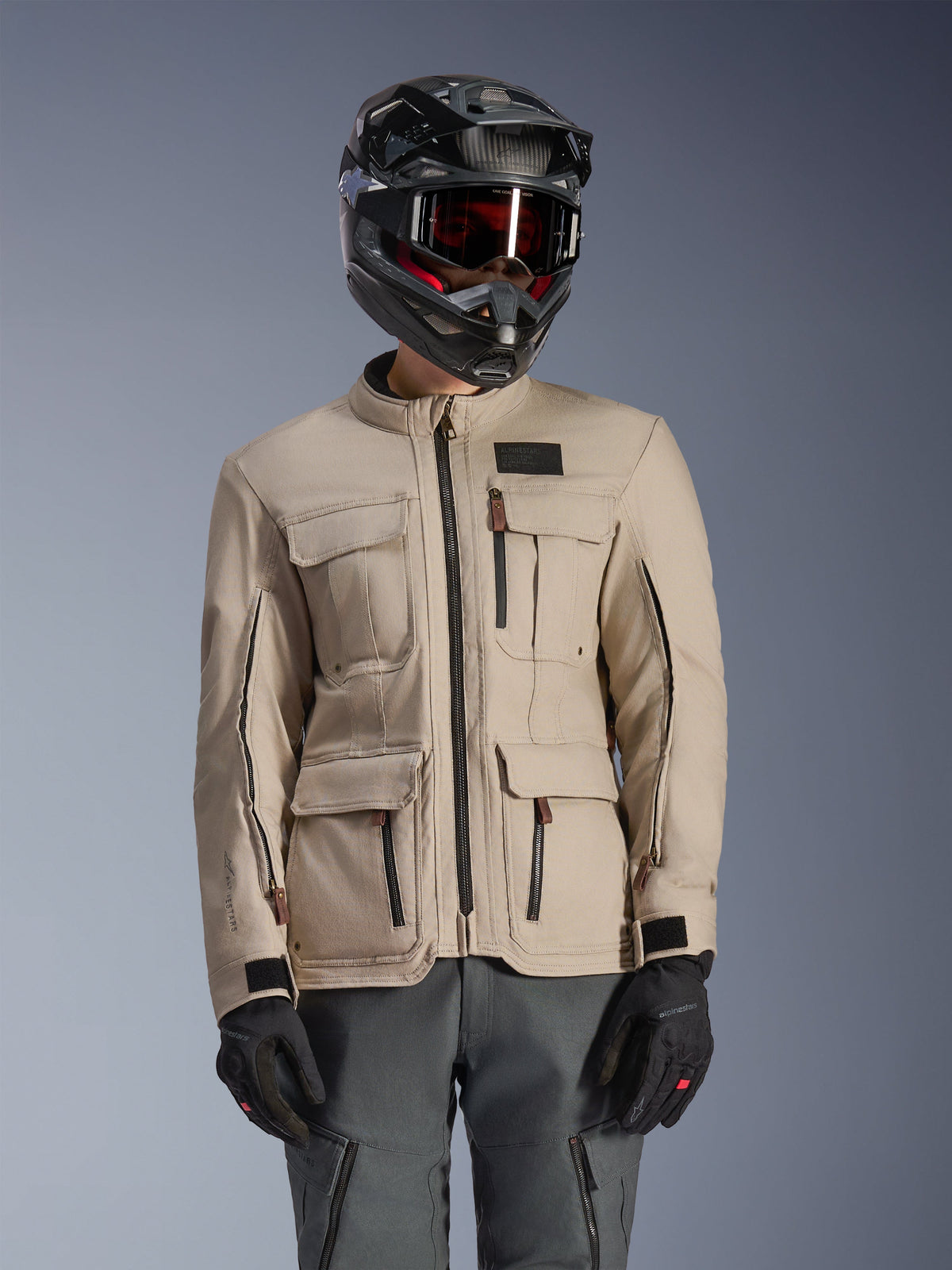 Alpinestars Borrego Drystar® Jacket, veste de moto d'aventure, gris chaud, dotée de multiples poches cargo à l'avant et de fermetures éclair noires, portée avec un casque noir, des lunettes et un pantalon de moto gris foncé, conception textile robuste pour le touring