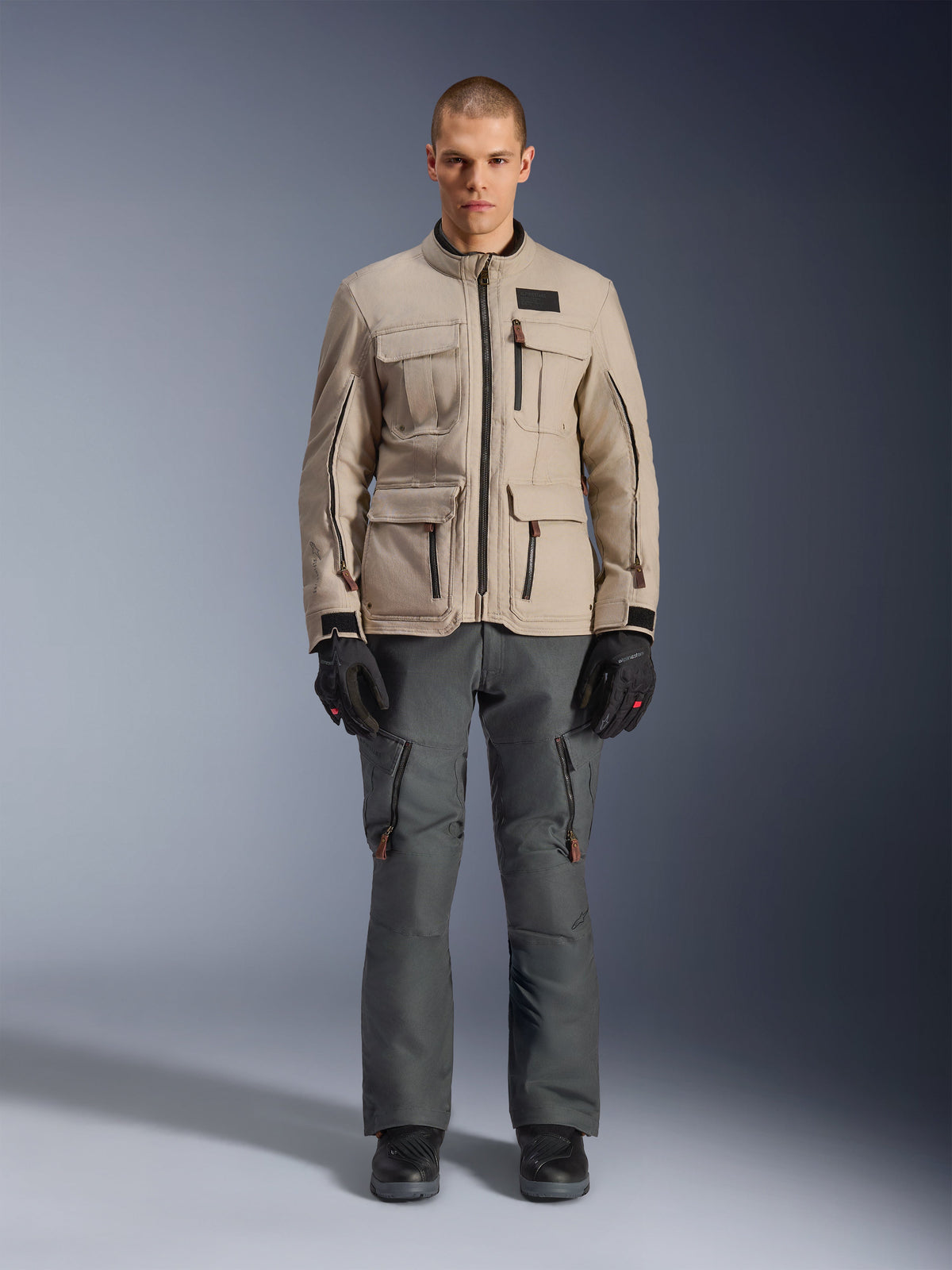 Veste Alpinestars Borrego Drystar®, veste de moto d'aventure, Warm Gray, portée par un mannequin avec un pantalon de moto gris foncé et des gants noirs, dotée de multiples poches cargo et d'une construction textile durable avec un aspect classique en toile cirée.