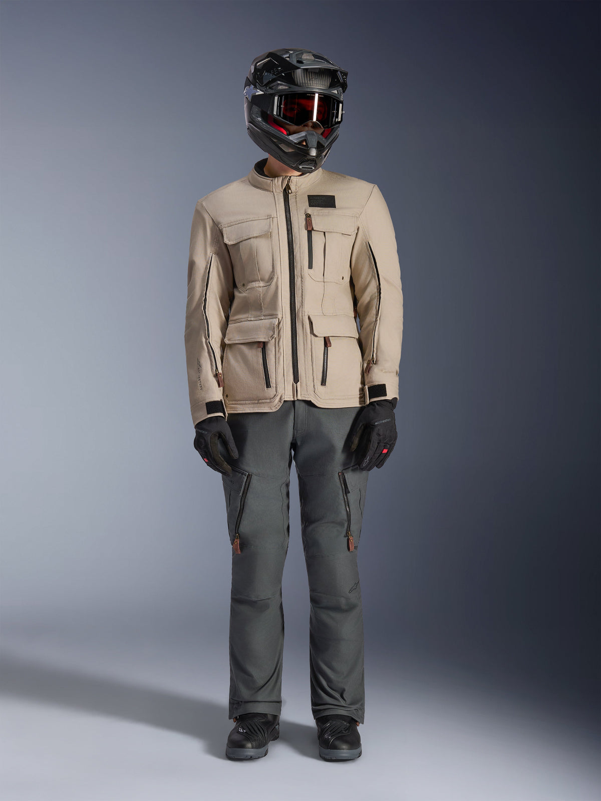 Veste Alpinestars Borrego Drystar® en Warm Gray, veste de moto d'aventure avec quatre poches cargo à l'avant, associée à un pantalon de moto gris foncé, un casque tout-terrain noir et des gants noirs, présentant un aspect toile cirée robuste