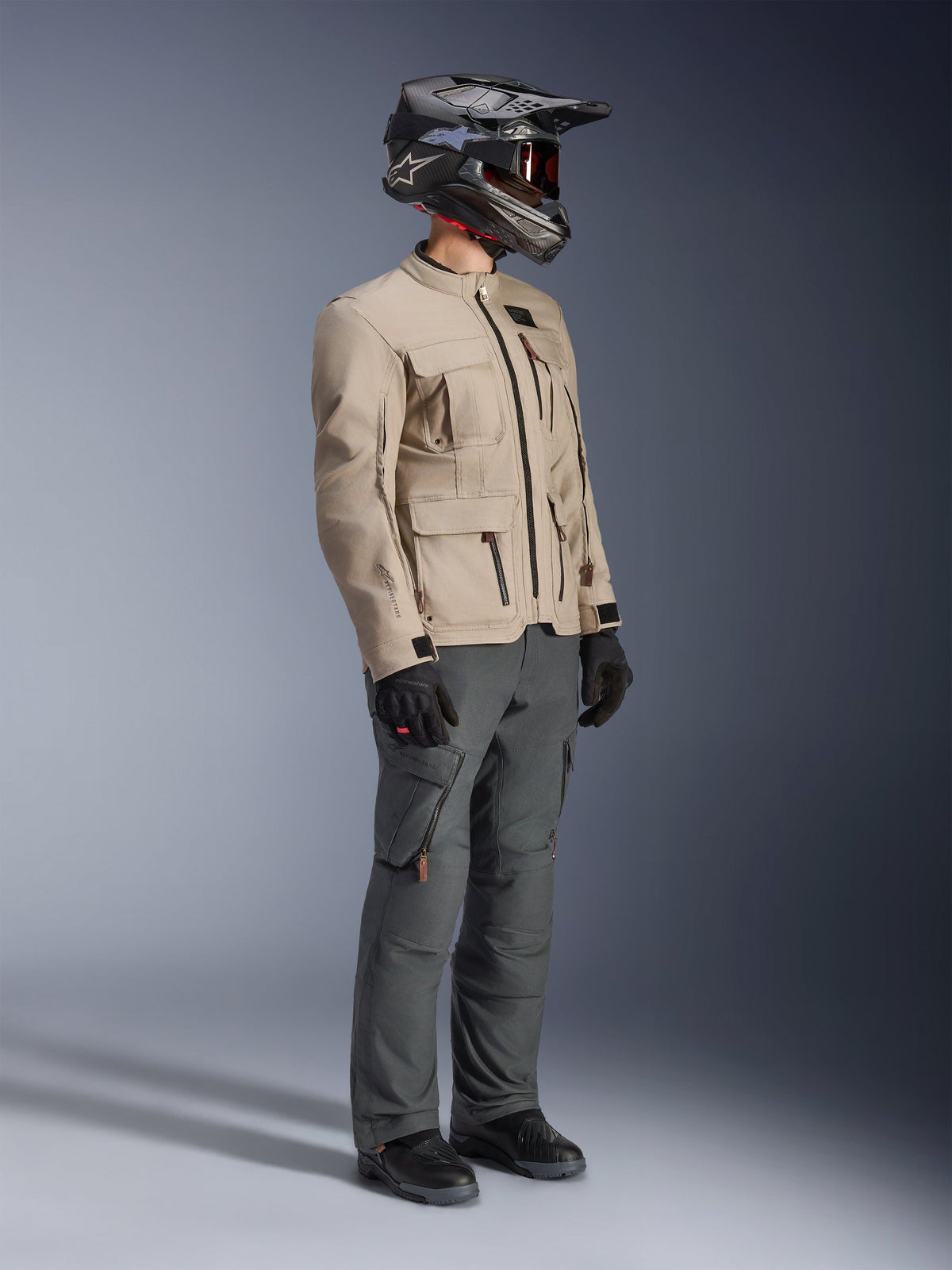 Motard portant la veste Alpinestars Borrego Drystar® en Warm Gray, une veste de moto d'aventure avec une texture en toile cirée et des poches utilitaires, associée à un pantalon de moto gris foncé, un casque en fibre de carbone, des gants noirs et des bottes noires, vue studio en pied d'une tenue de touring aventure complète.