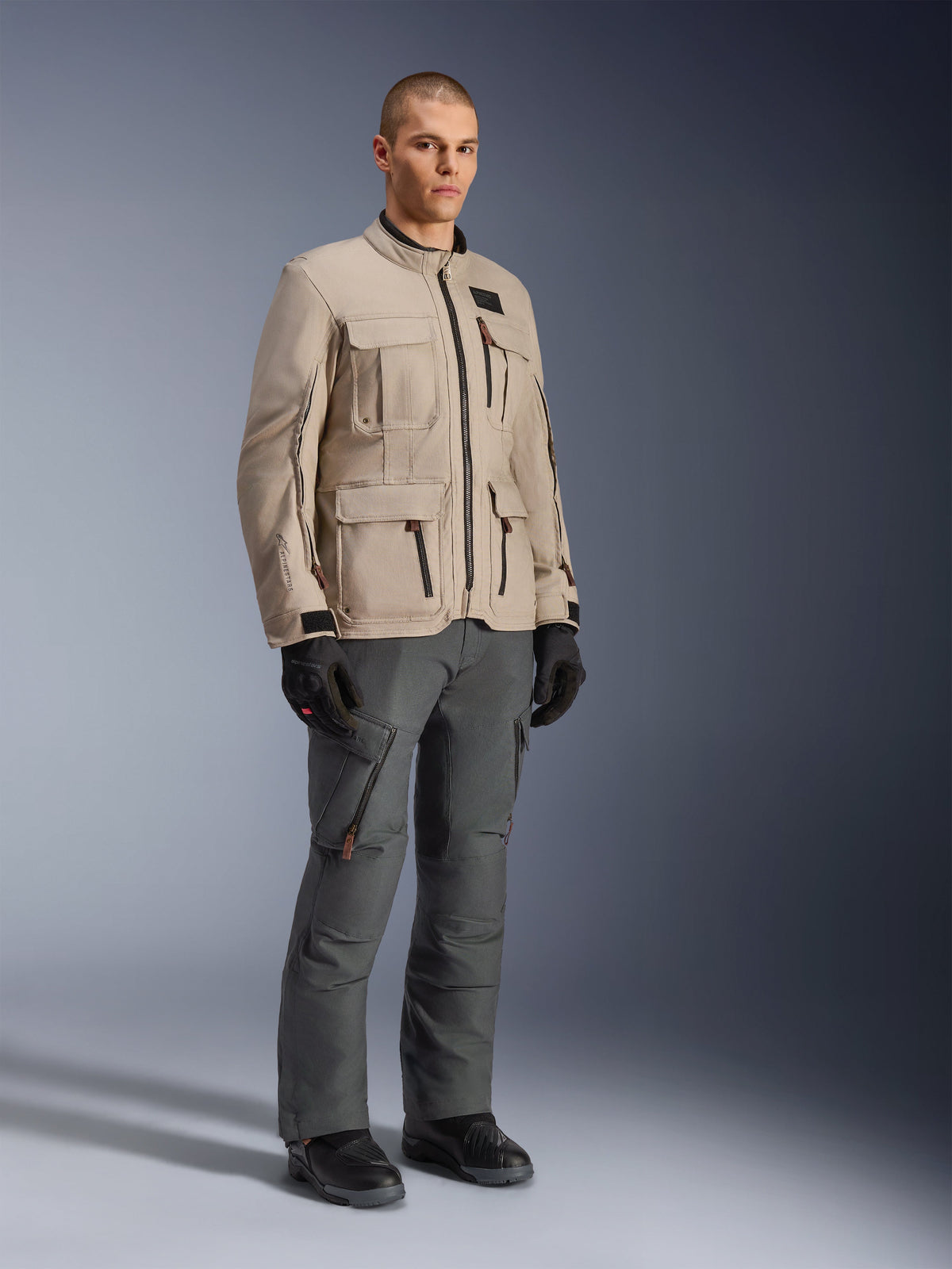 Veste Alpinestars Borrego Drystar® en Warm Gray, veste de moto d'aventure avec un aspect toile cirée et de multiples poches utilitaires, portée par un mannequin avec un pantalon de moto gris foncé, des gants noirs et des bottes noires, portrait de studio en pied