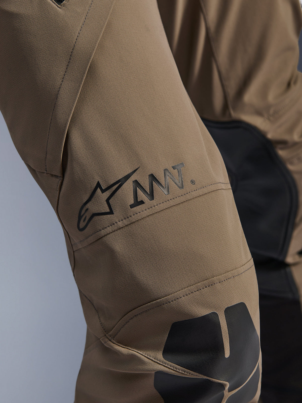Pantalon Alpinestars AMT-8 Stretch Drystar® XF en Walnut Black, gros plan sur la jambe présentant le logo AMT noir et l'étoile Alpinestars, textile extensible haute performance avec empiècements noirs, équipement technique de moto d'aventure
